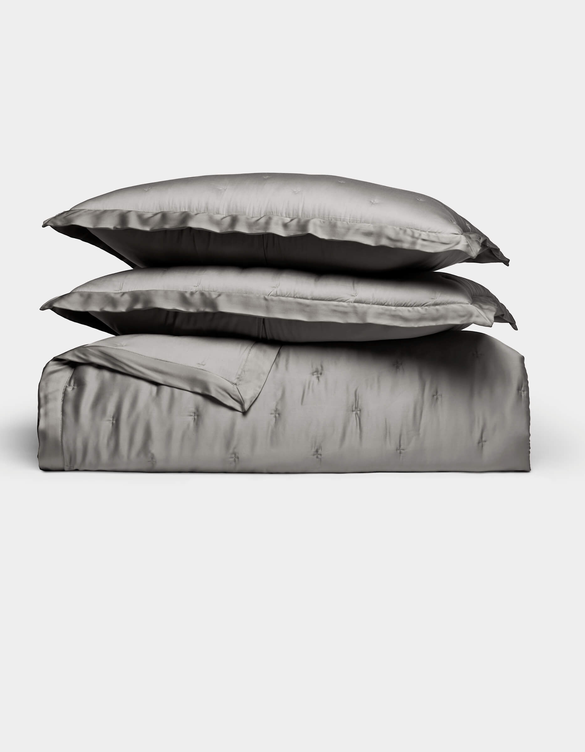 The Bamboo Quilt Bundle by Cozy Earth features lustrous silver-gray bed linens, including two stacked pillows, a folded sheet, and a duvet cover, all arranged against a plain white background.|Color:Charcoal