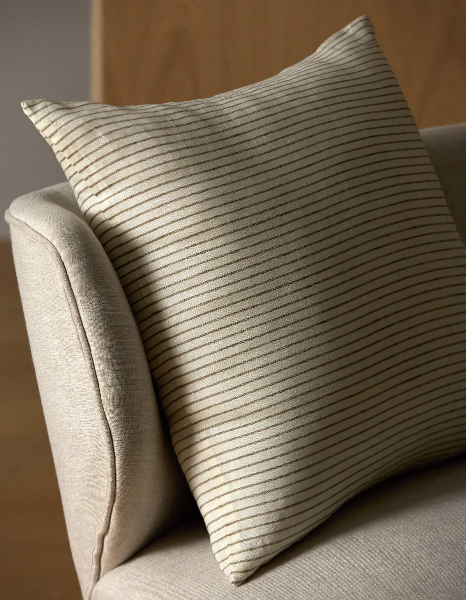 A close-up of the Cozy Earth Striped Throw Pillow resting on a light armchair, with a blurred ceramic vase and branches in the background.|Color:Narrow Stripe Sable and Coconut |Size:26" x 26"