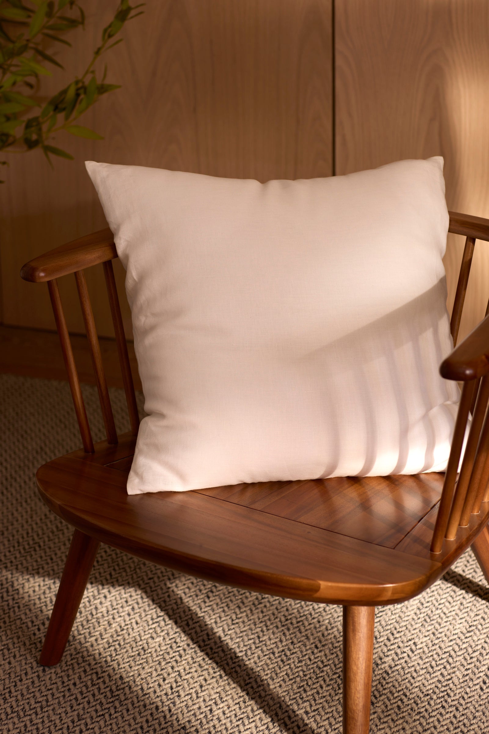 A Cozy Earth Linen Throw Pillow rests on a wooden chair with wide, angled arms, placed on a textured rug. Soft natural light casts shadows across the white pillow and wood.|Color:White |Size:20" x 20"