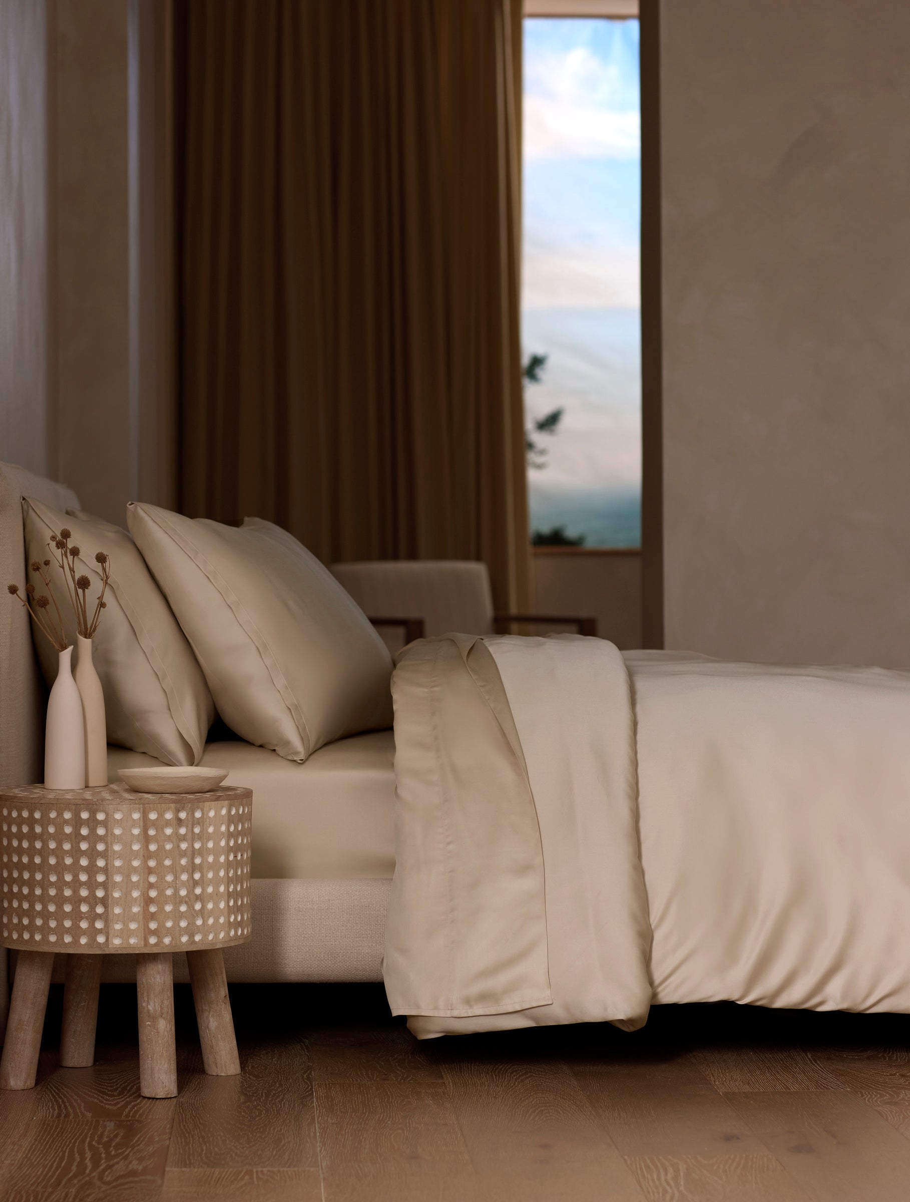 A modern bedroom features a neatly made bed with Cozy Earth’s Bamboo Sheet Set in cream, a wooden side table with dried flowers, neutral walls, wooden flooring, and a window revealing a blue sky with clouds.|Color:Driftwood