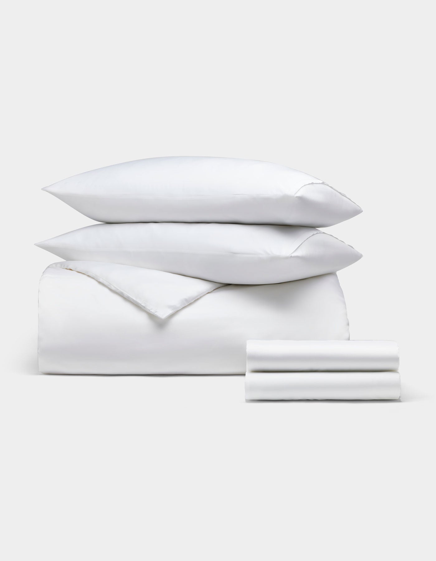 The Bamboo Bedding Deluxe Bundle by Cozy Earth features a neatly stacked set of white bedding—two pillows, a comforter, and a folded sheet—displayed against a plain light gray background.|Color:White