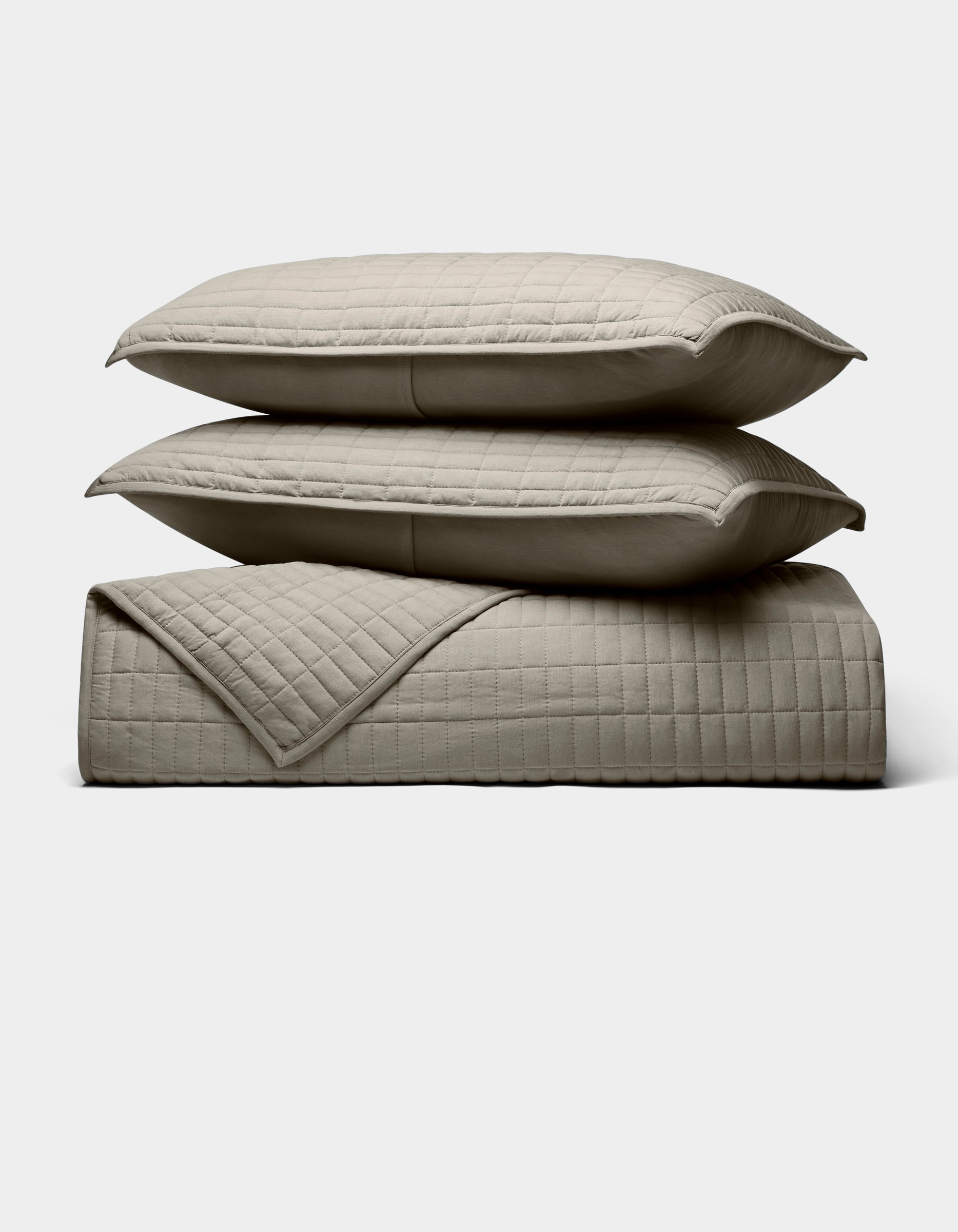 Two beige Cozy Earth Bamboo Jersey Quilt Bundle pillows are stacked atop a matching quilted blanket, all neatly arranged against a plain light background.|Color:Stone