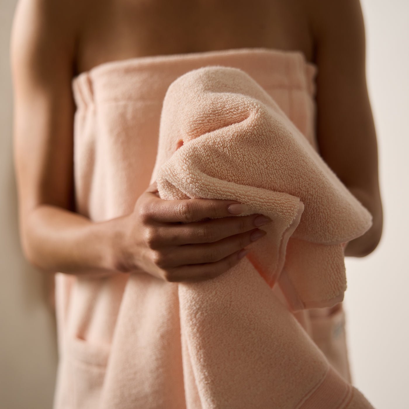A person holds a soft, peach-colored Luxe Bath Sheet from Cozy Earth against their chest, while wearing another matching towel wrapped around their body. The background is neutral and out of focus. |Color:Blossom