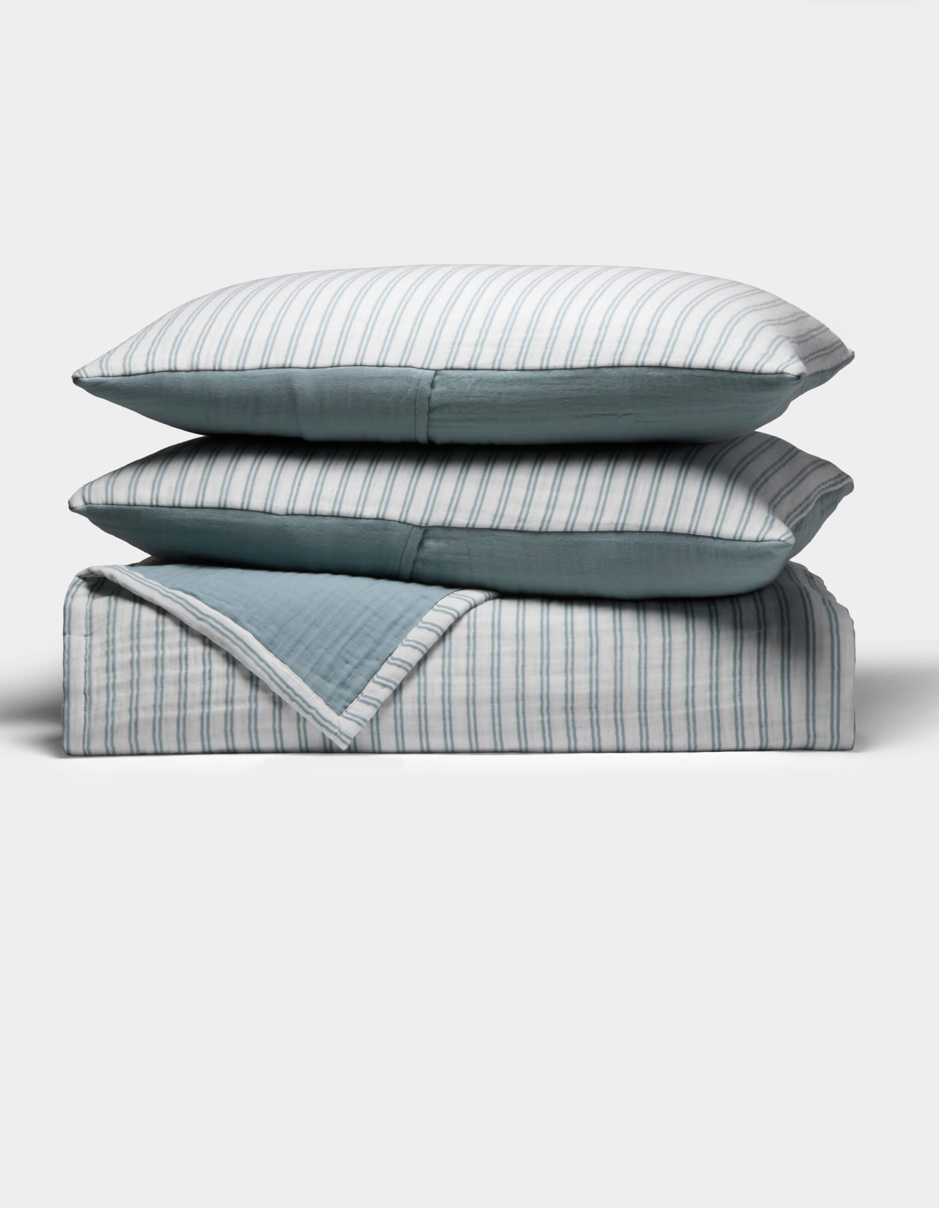 The Cozy Earth Waterfront Coverlet Bundle shows two pillows and a folded quilt stacked neatly, each with blue and white stripes on one side and solid blue on the other, all set against a plain white background.|Color:River Stripe