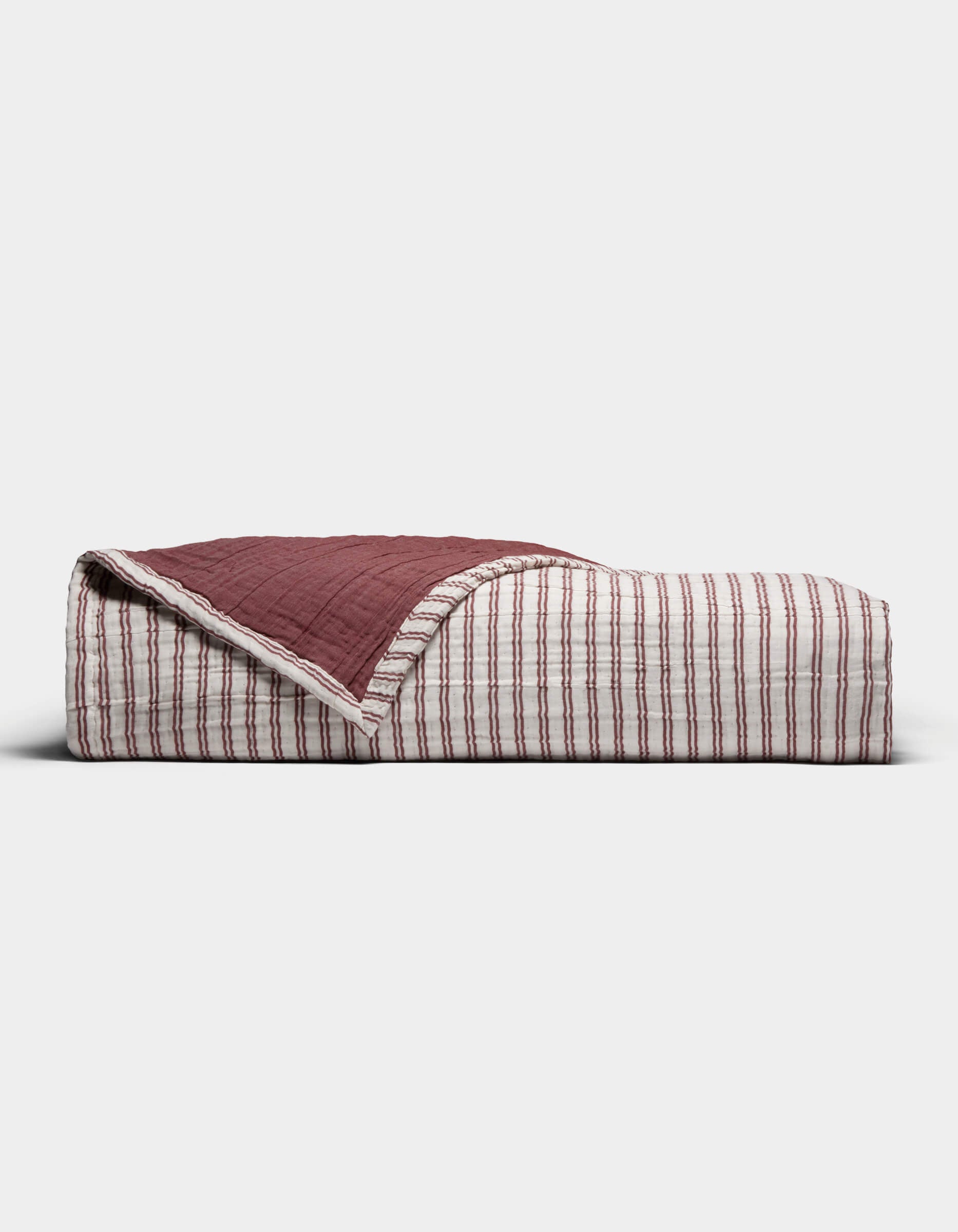 The Cozy Earth Waterfront Coverlet, a folded quilt in red and white stripes with one corner flipped to reveal a solid dark red underside, is displayed against a plain white background.|Color:Currant Stripe