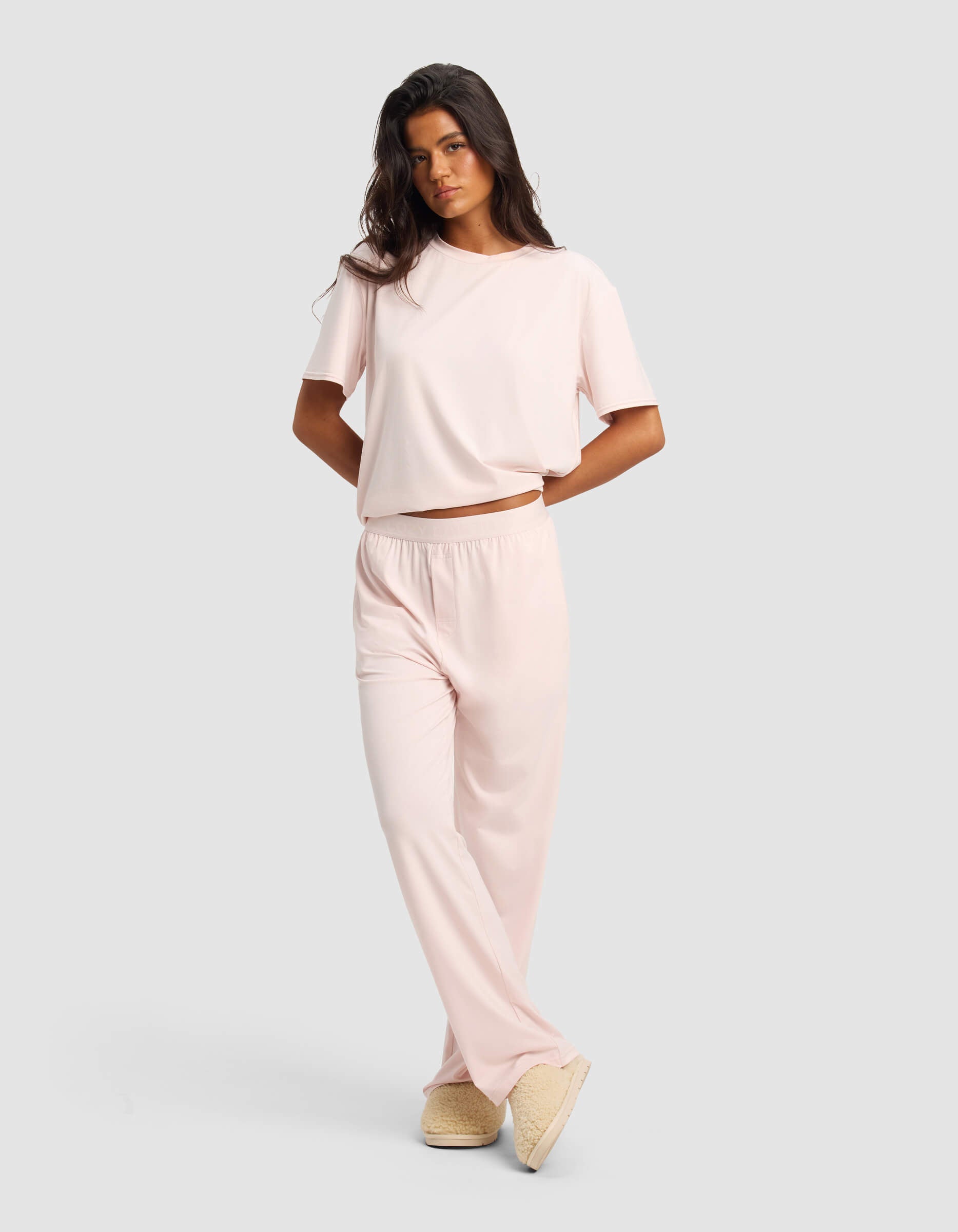A woman models the Cozy Earth Women's First Layer Pajama Short Sleeve Tee & Pant Set in light pink with beige slippers, standing against a plain light gray background.|Color:Ballet