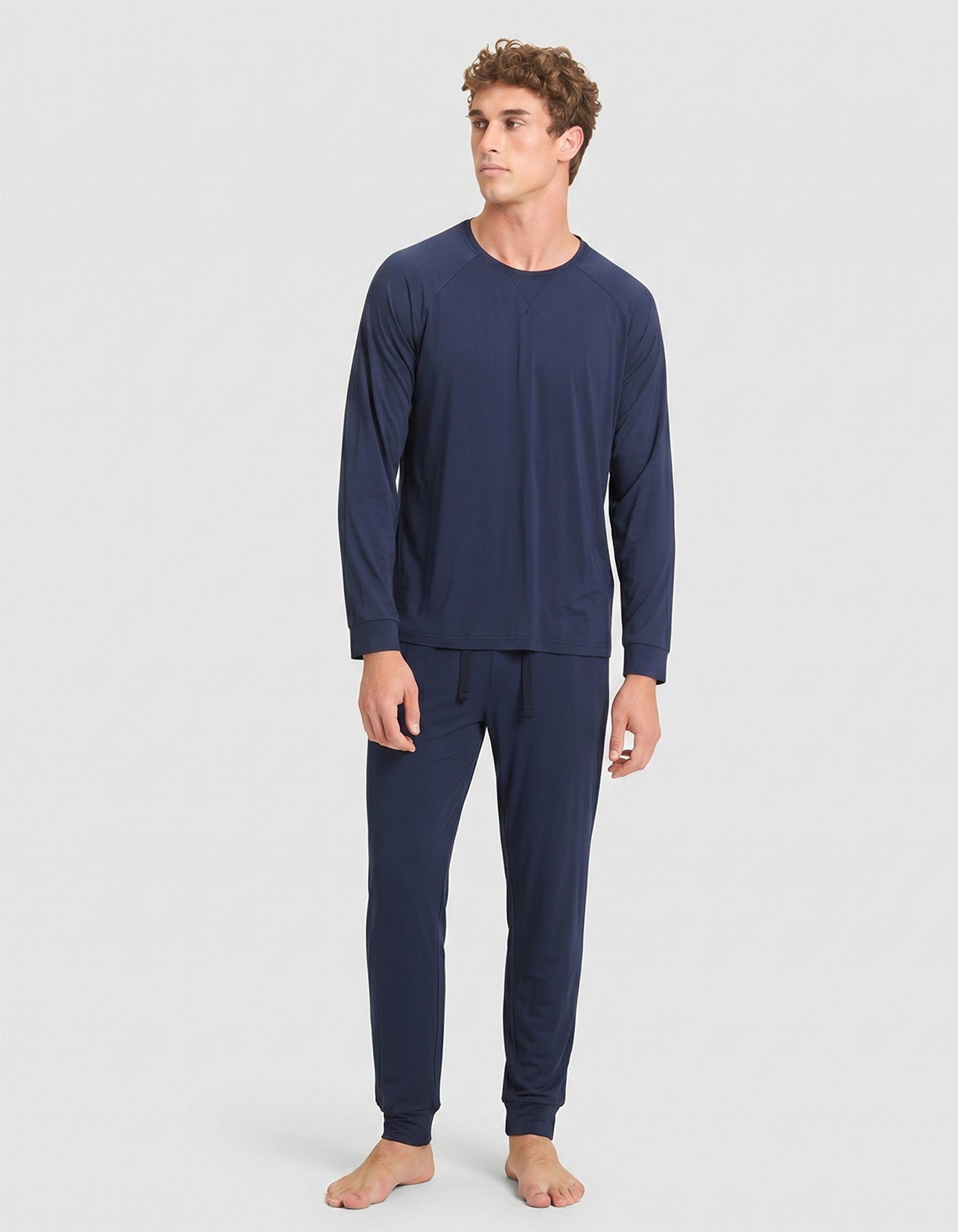 A young man with curly hair wears Cozy Earth's Men's Bamboo Stretch-Knit Long Sleeve Pajama Set in navy blue, standing against a plain light gray background and looking slightly to his left. |Color:Navy