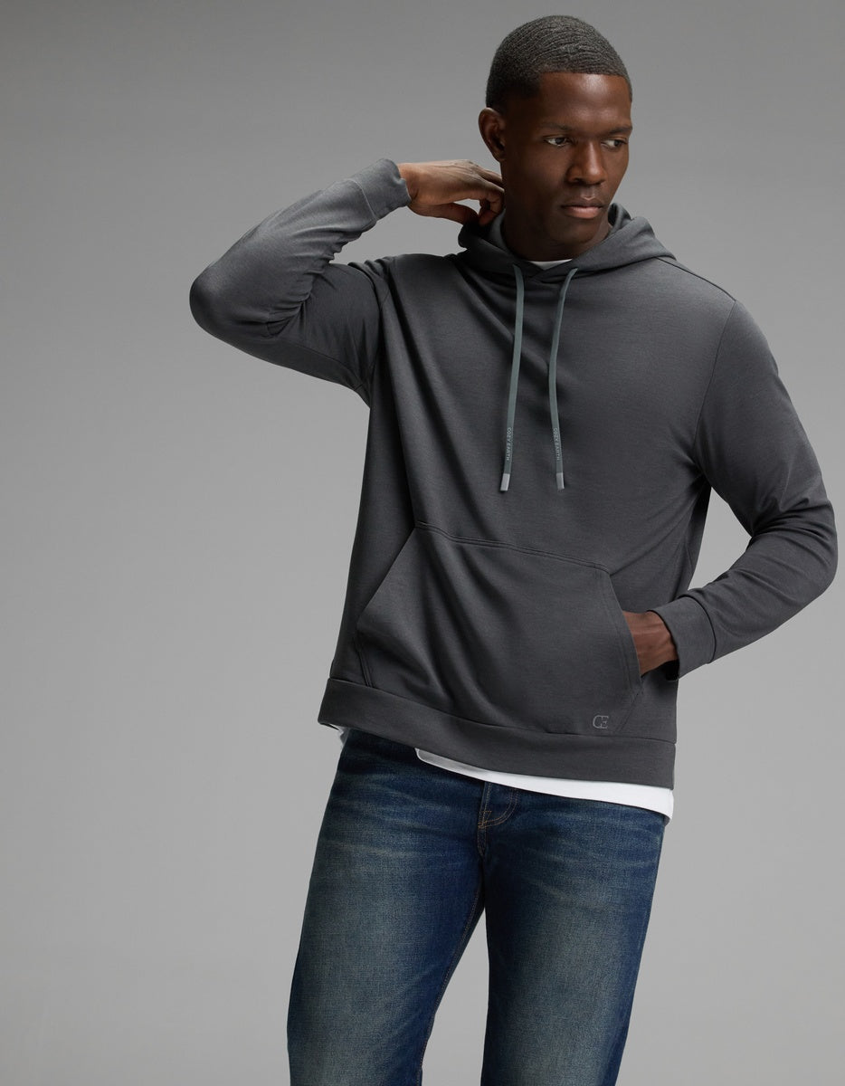 A man models the Cozy Earth Men’s Brushed Bamboo Hoodie in dark gray with blue jeans, standing against a plain gray background and gazing to the side while resting one hand on the back of his neck.|Color:Charcoal