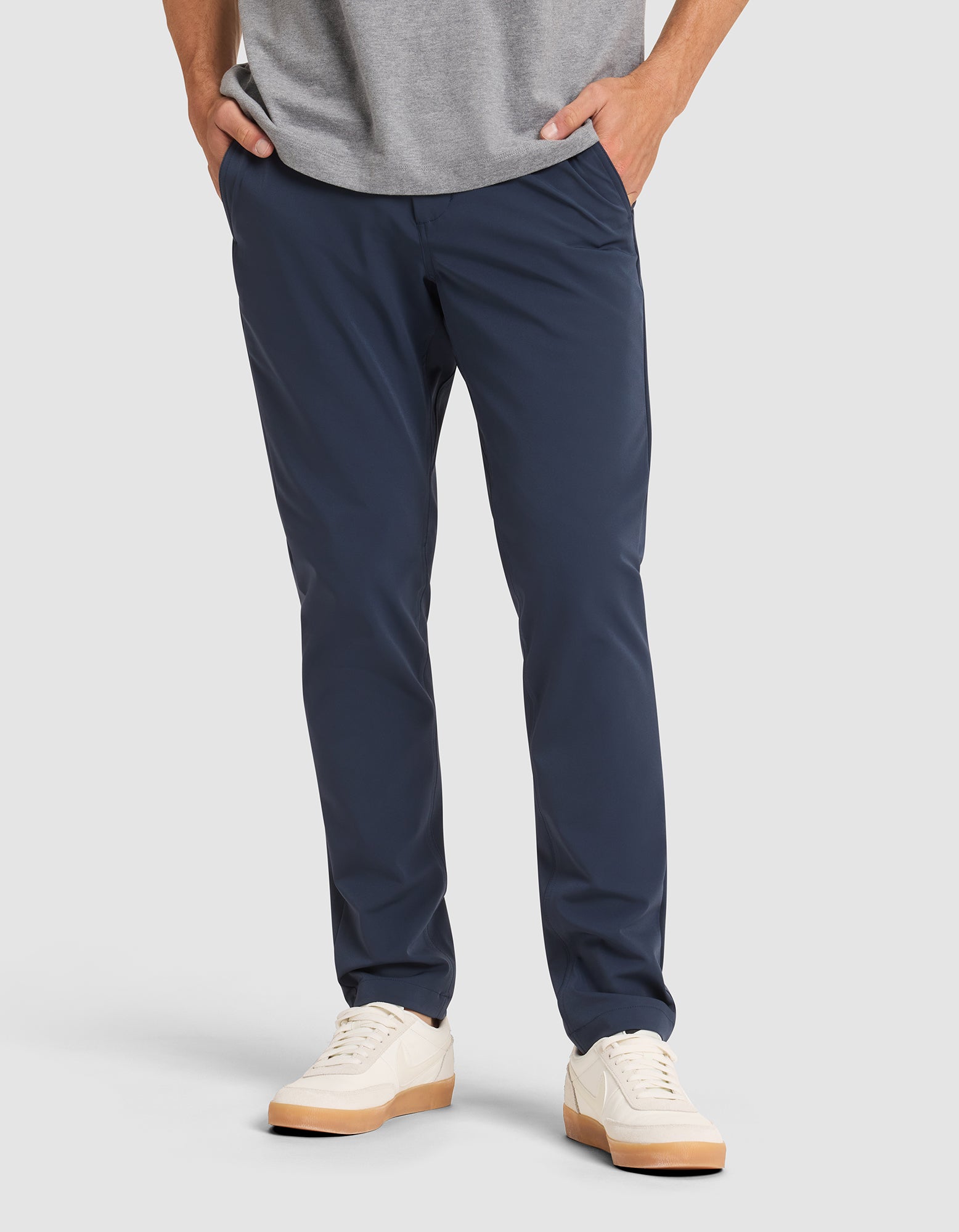 A person stands with hands in pockets against a plain background, wearing Cozy Earth's Men's Everywhere Pant in navy blue, a gray shirt, and white sneakers with light brown soles. |Color:Eclipse