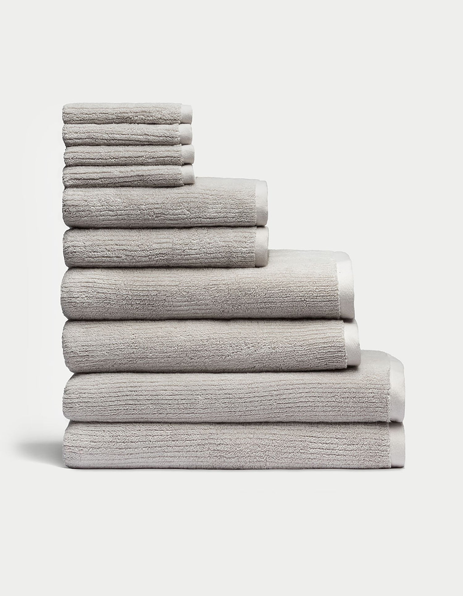 The Complete Ribbed Terry Bath Bundle by Cozy Earth features a neatly stacked arrangement of plush, gray towels in various sizes, piled eight layers high against a plain white background, showcasing their soft and textured surface for ultimate comfort.|Color:Light Grey