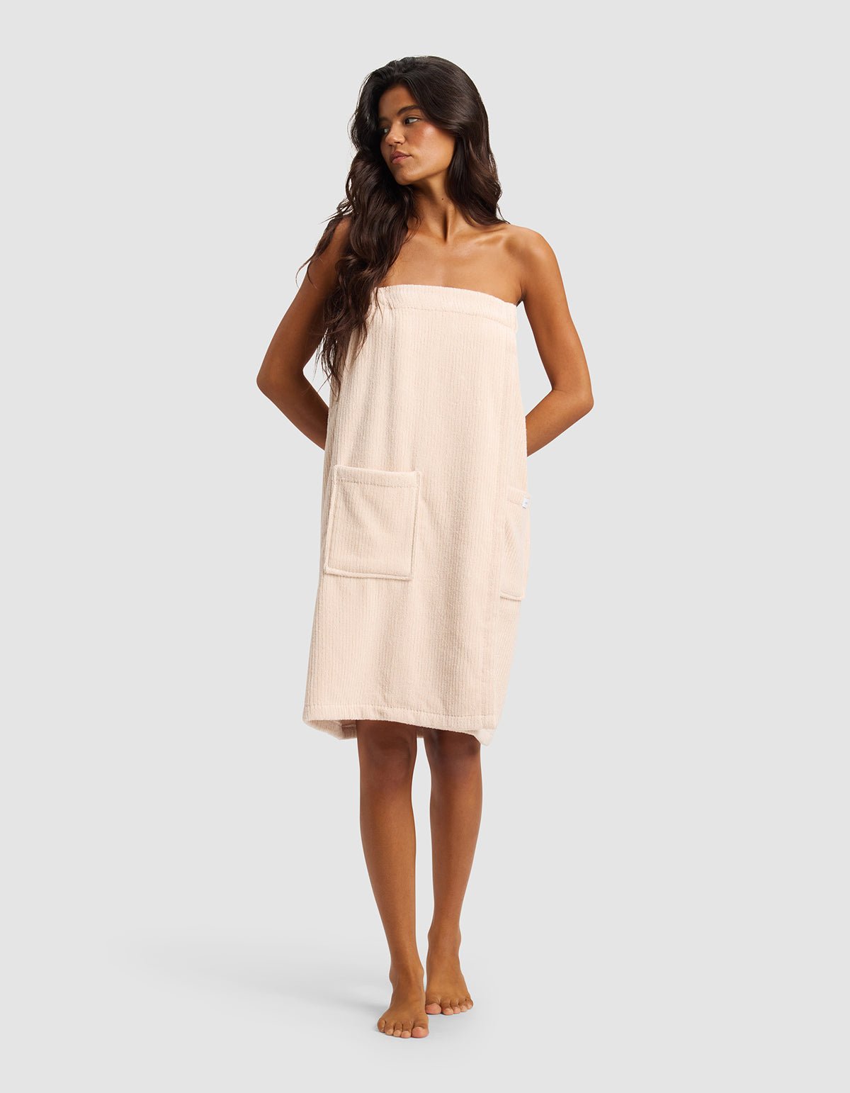 A woman with long dark hair stands barefoot against a plain light gray background, wearing the Cozy Earth Ribbed Terry Bath Wrap in light beige with two front pockets.|Color:Petal