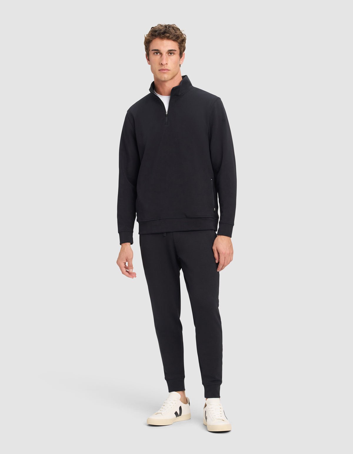 A man stands against a plain background wearing Cozy Earth's Men's StretchTech Quarter Zip in black, matching black joggers, and white sneakers. He faces forward with a neutral expression and relaxed posture.|Color:Jet Black
