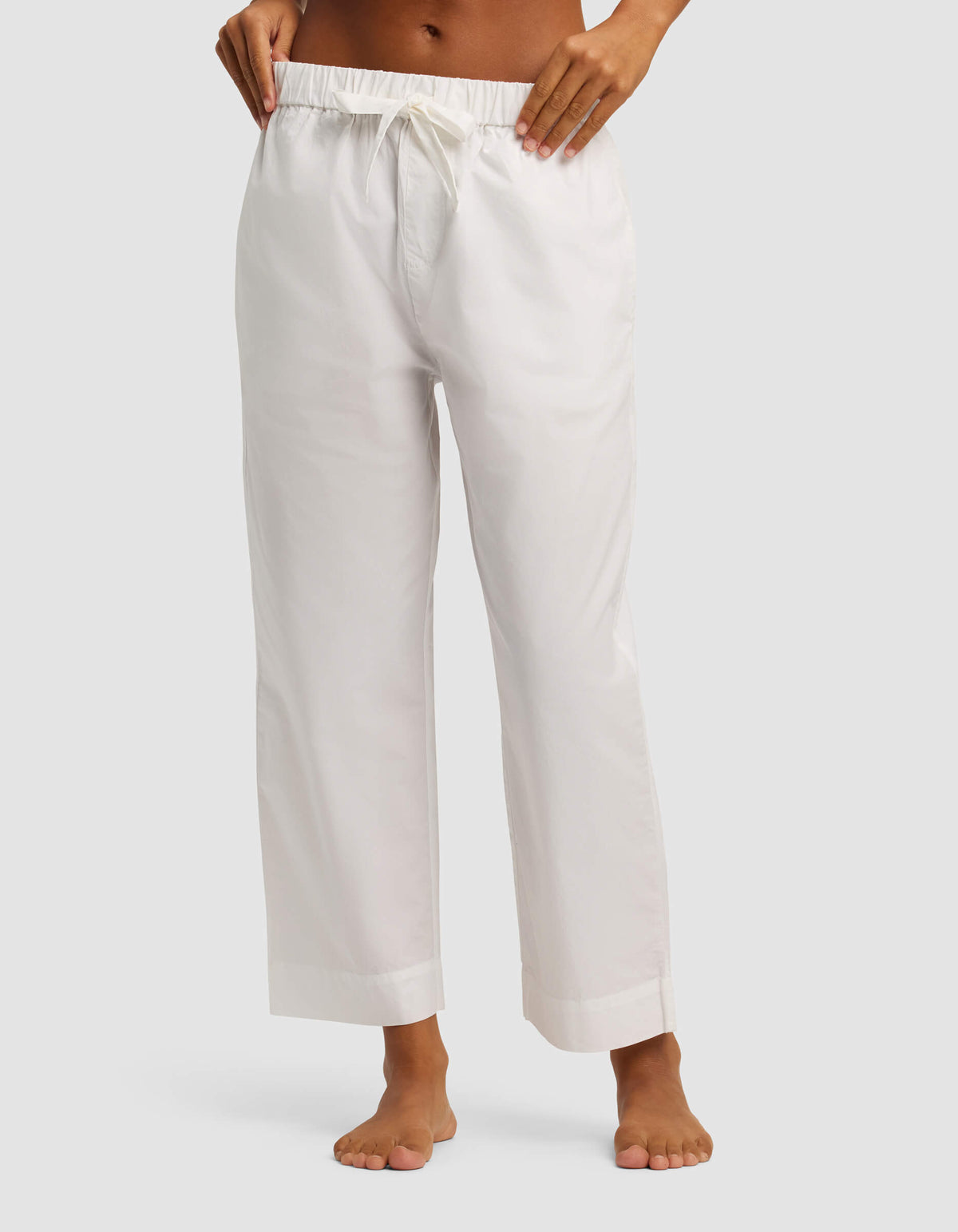 A person stands barefoot against a plain light gray background, wearing Cozy Earth’s Women's Sutton Cropped Pajama Pant—loose-fitting with a white drawstring and cropped just above the ankles.|Color:White