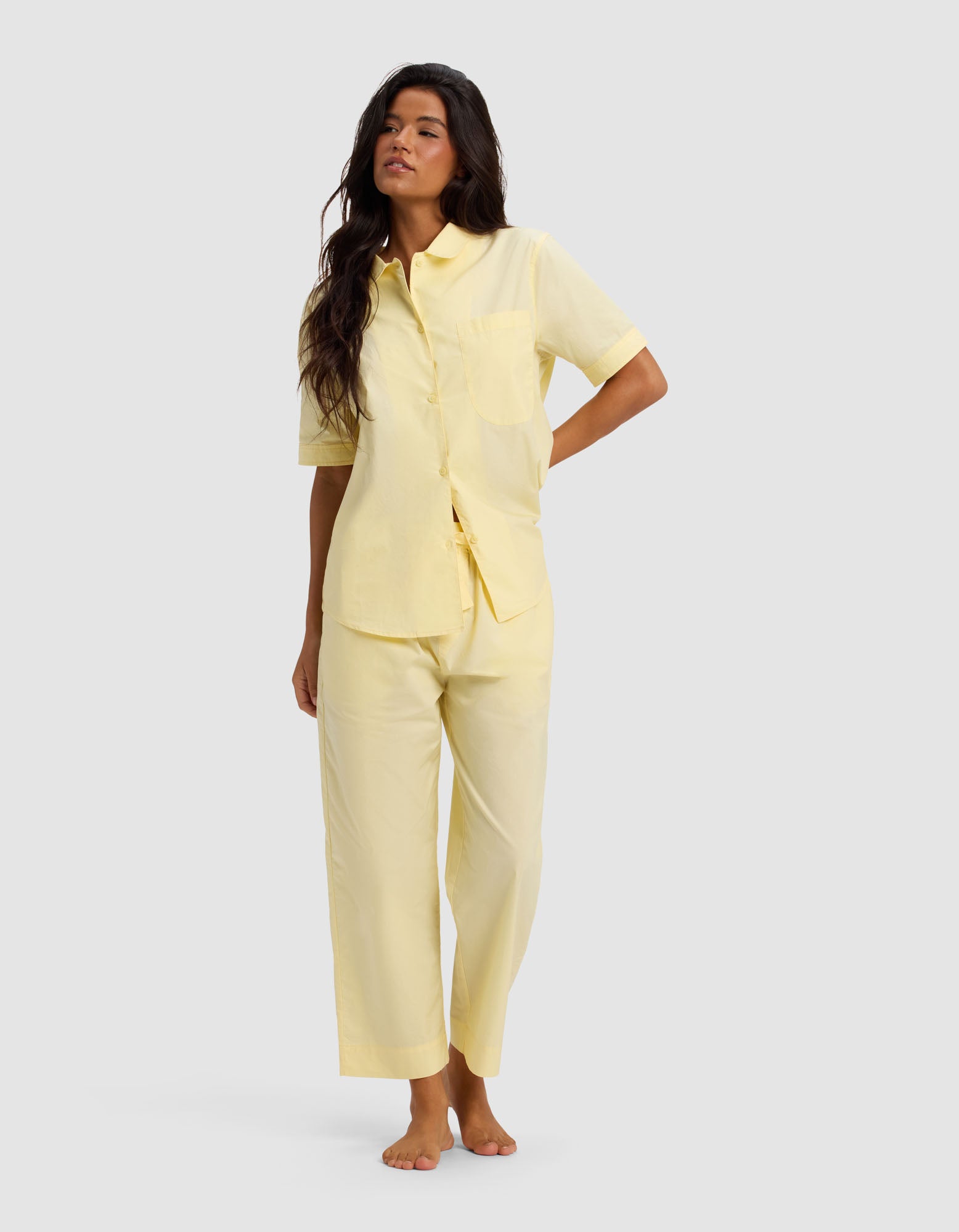 A woman with long, wavy dark hair stands barefoot against a light gray background, wearing the Cozy Earth Women's Sutton Short Sleeve & Cropped Pant Pajama Set in light yellow. She looks relaxed.|Color:Meringue