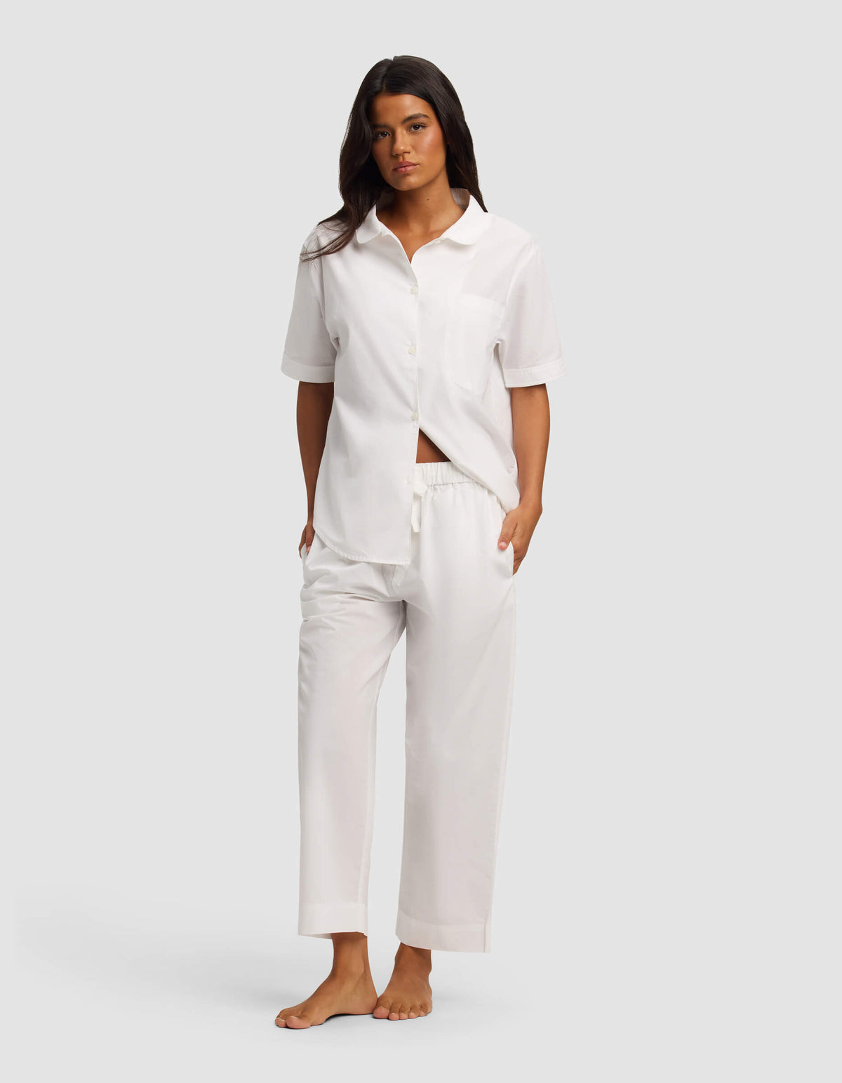 A woman stands barefoot against a plain background, wearing the Cozy Earth Women's Sutton Short Sleeve & Pant Pajama Set in white. The loose-fitting set features a button-up shirt, short sleeves, and matching drawstring pants. Her hands are in her pockets.|Color:White