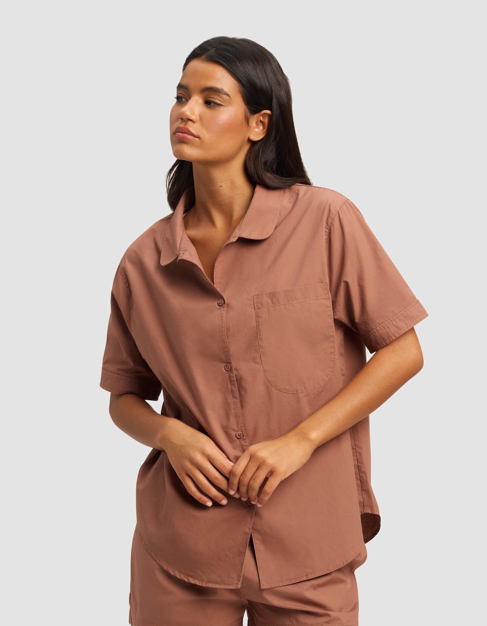A woman with long dark hair wears the Cozy Earth Women's Sutton Short Sleeve Pajama Top and matching shorts in warm brown, standing with one hand resting on the other against a plain light gray background.|Color:Saddle