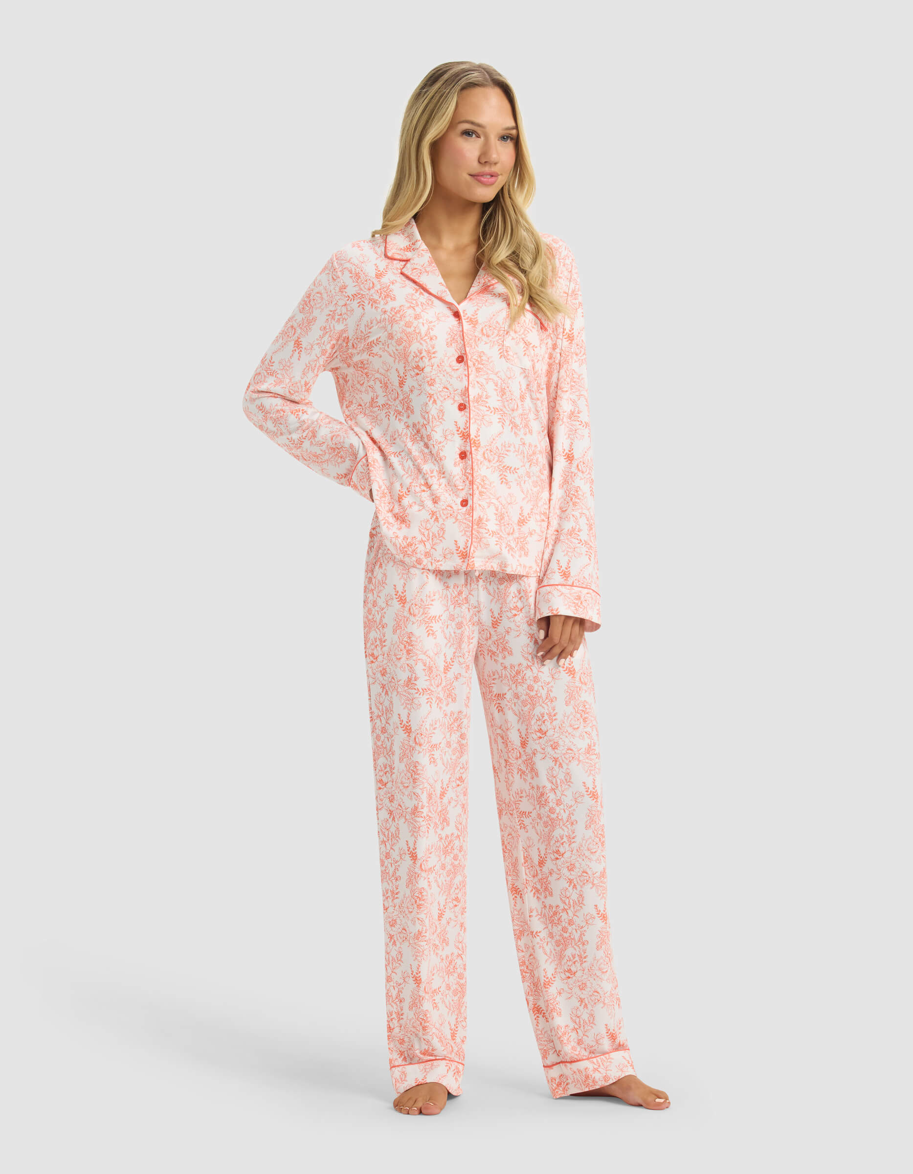 A woman stands barefoot in Cozy Earth’s Women's Bamboo Stretch-Knit Long Sleeve Pajama Set, featuring a pink and white pattern with a button-up top and matching pants, against a plain light gray background.|Color:Melon Toile