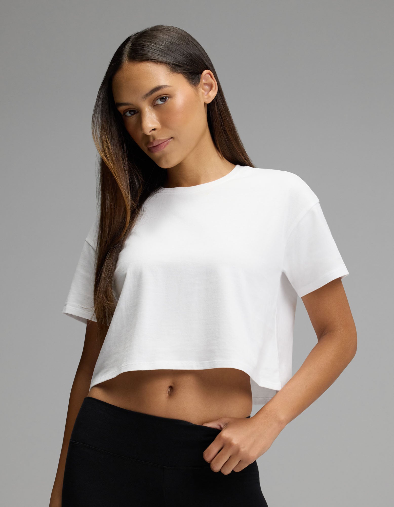 A woman with long brown hair stands confidently against a plain gray background, wearing the Cozy Earth Women’s All Day Cropped Tee in white paired with black pants.|Color:White