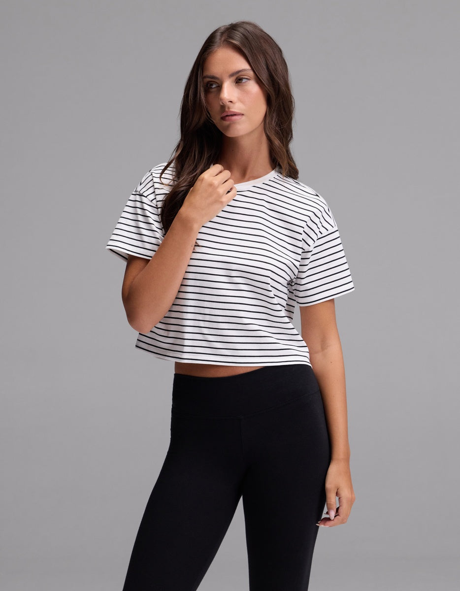 A woman with long brown hair is wearing the Cozy Earth Women’s All Day Cropped Tee in Jet Black Stripe and black leggings, standing against a plain gray background.|Color:Jet Black Stripe