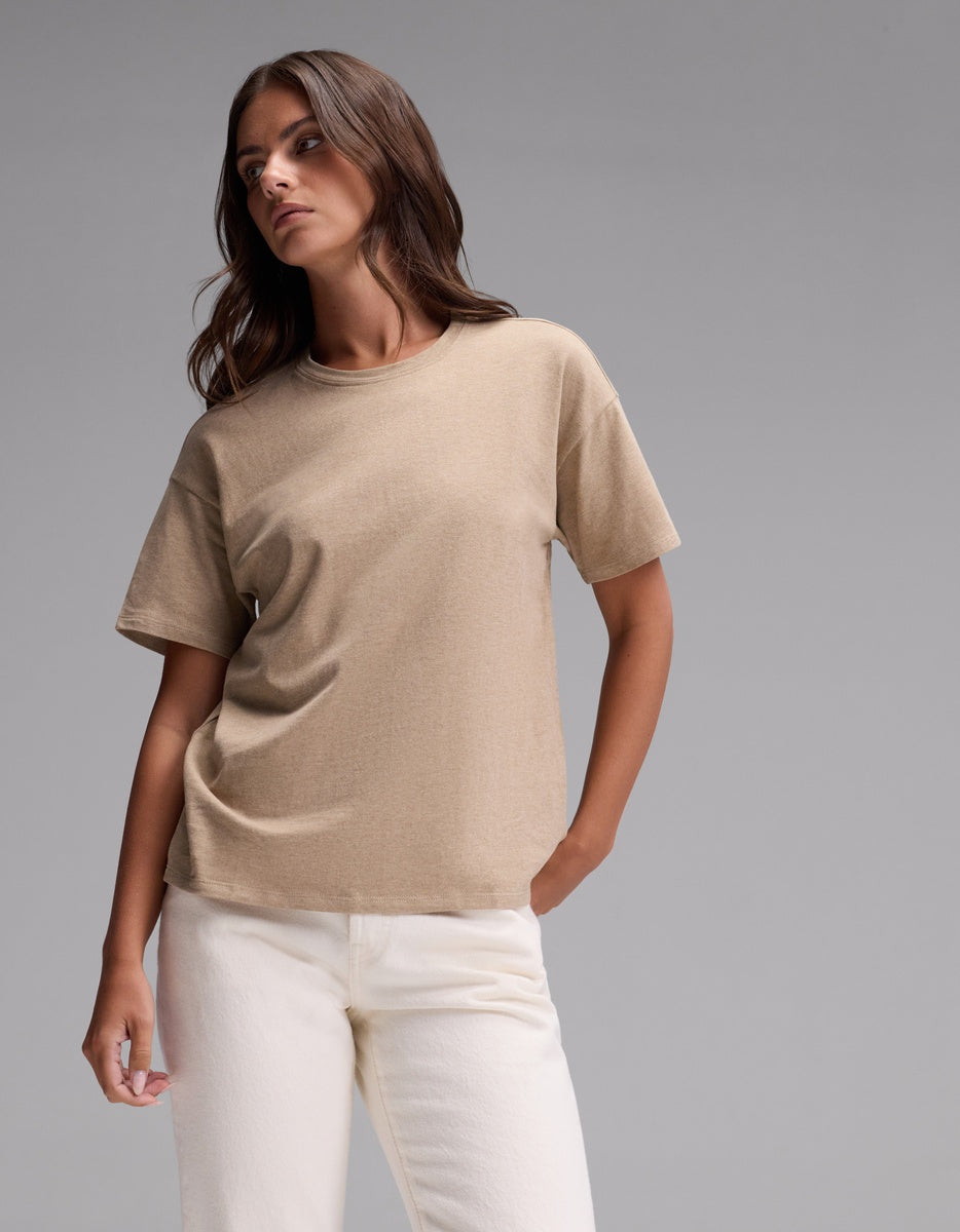 A person stands against a plain gray background, wearing the Cozy Earth Women’s All Day Tee in Beech Heather paired with white pants. They have long brown hair and a relaxed pose, looking slightly to the side.|Color:Beech Heather