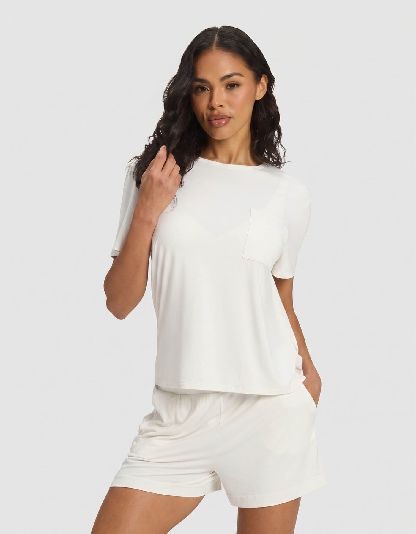A woman wearing the Cozy Earth Women's Bamboo Stretch-Knit Lounge Tee in Ivory stands against a plain light background, pairing the top with matching shorts, one hand in her pocket and the other touching her hair.|Color:Ivory