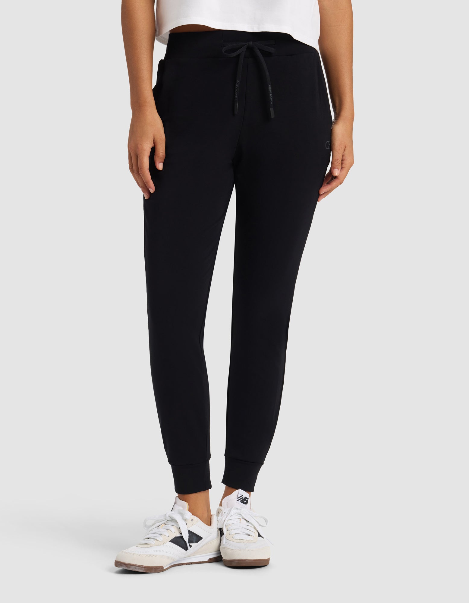 A person stands against a light gray background with hands in pockets, wearing Cozy Earth's Women's Brushed Bamboo Jogger Pant in black, paired with a white shirt and white sneakers.|Color:Black