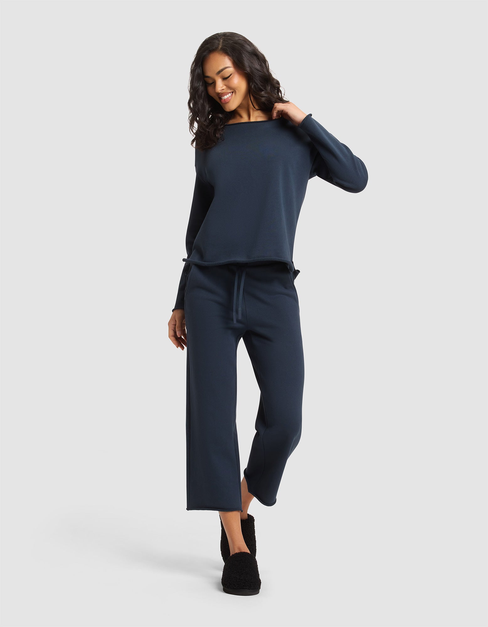 A woman with curly hair smiles and looks down while wearing Cozy Earth's Women's Chelsea Set—a navy blue long-sleeve top with matching wide-leg pants. She stands against a light gray background and wears black slippers. |Color:Eclipse