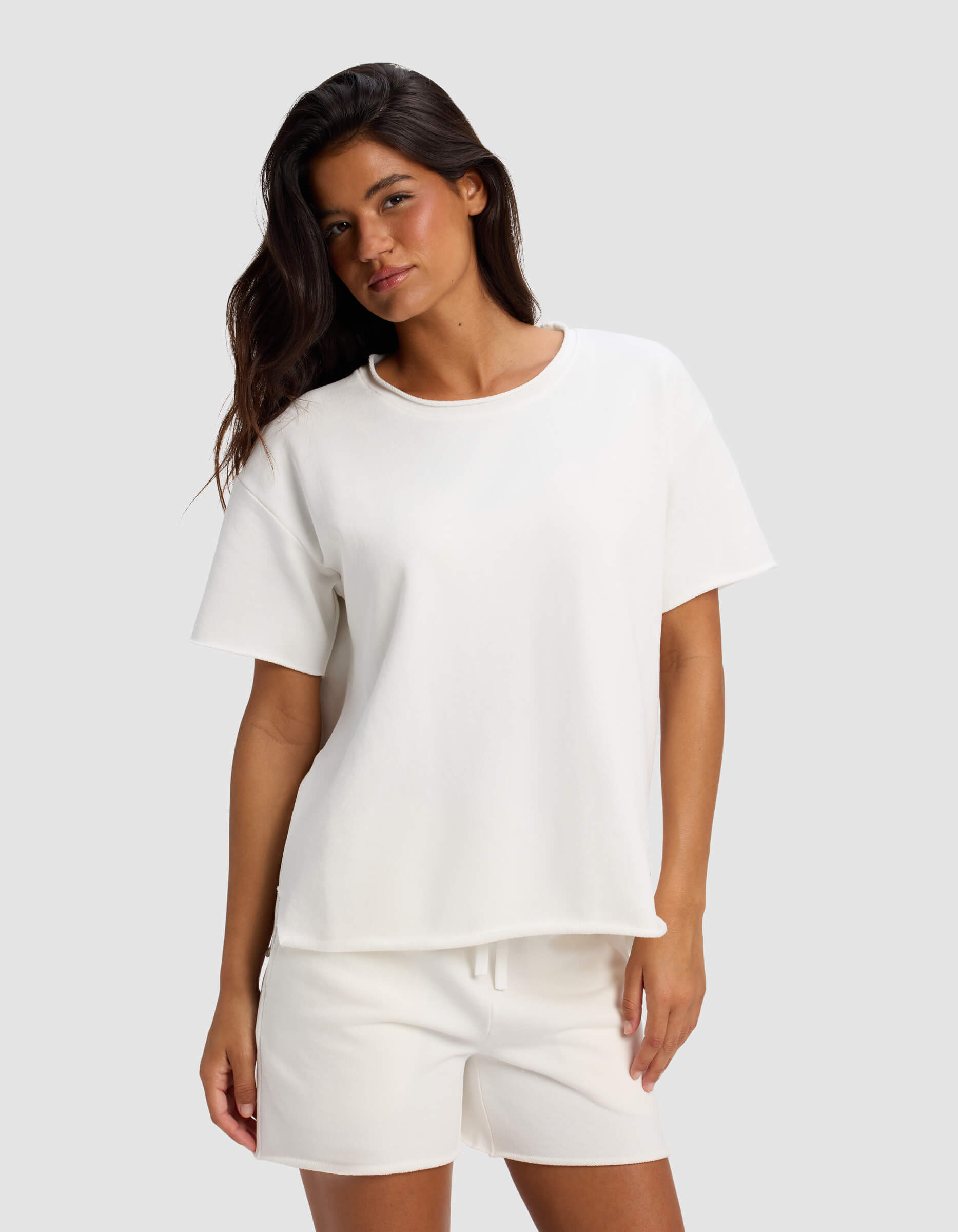 A woman with long dark hair models the Women's Chelsea Short Sleeve by Cozy Earth, paired with white shorts. She stands against a light gray background in a relaxed, casual pose with a slight smile.|Color:Bone