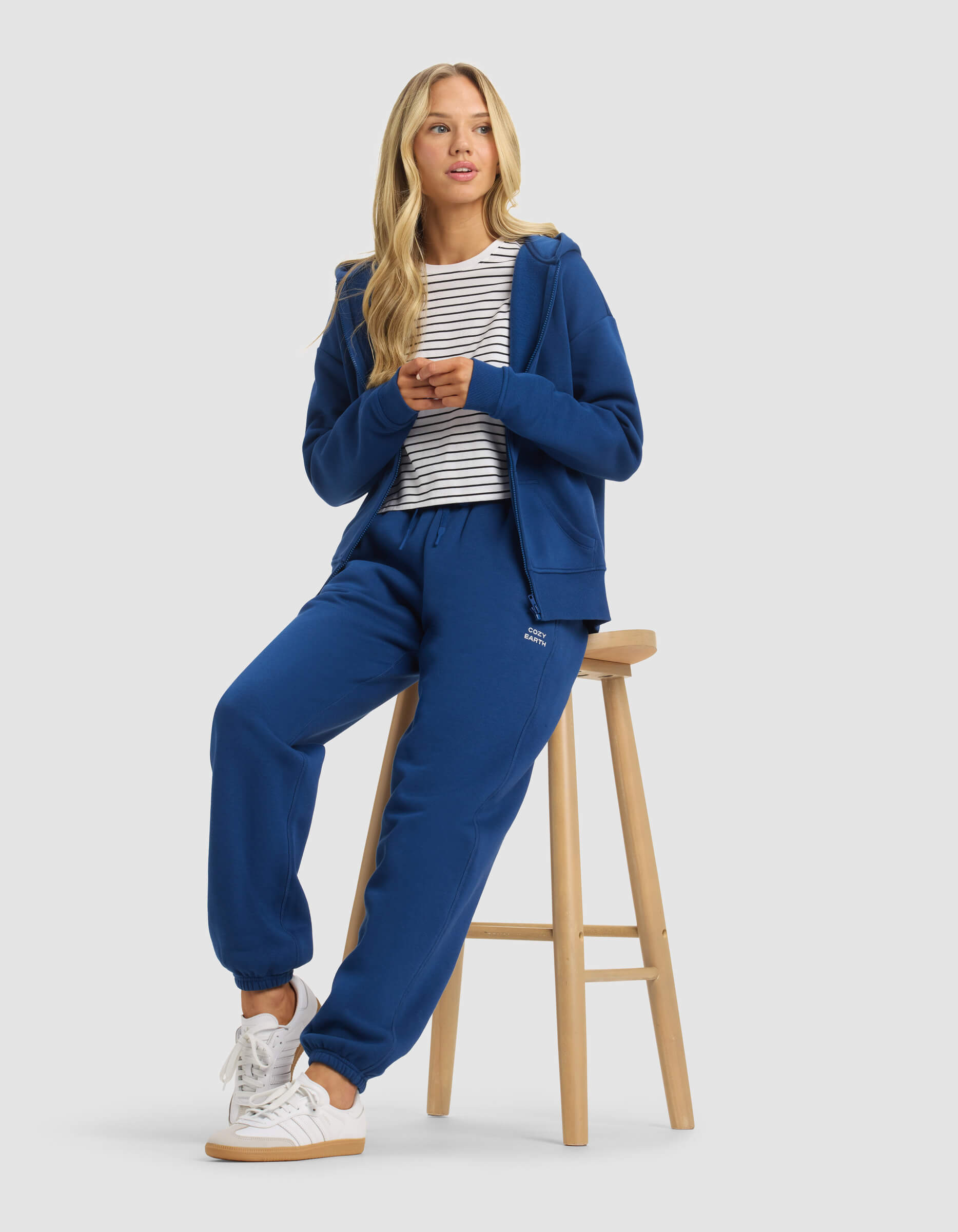 A woman with long blonde hair wears Cozy Earth's Women's Cityscape 2.0 Sweatpant in blue, paired with a white and black striped shirt and white sneakers. She sits on a wooden stool, hands together, looking slightly to the side.|Color:Pacific