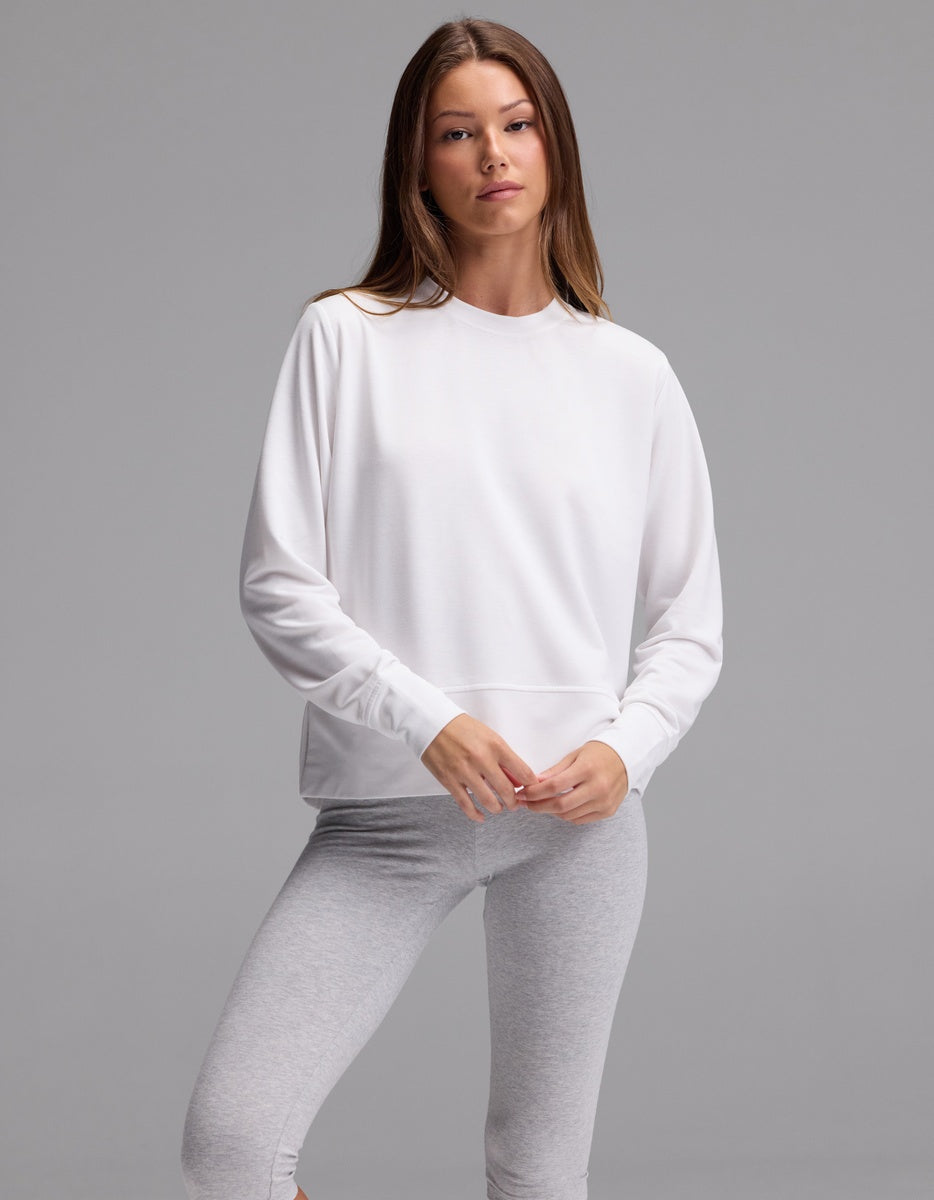 A woman with straight brown hair and a neutral expression stands against a plain gray background, wearing the Cozy Earth Women’s StudioLite Crewneck in white and light gray leggings.|Color:White