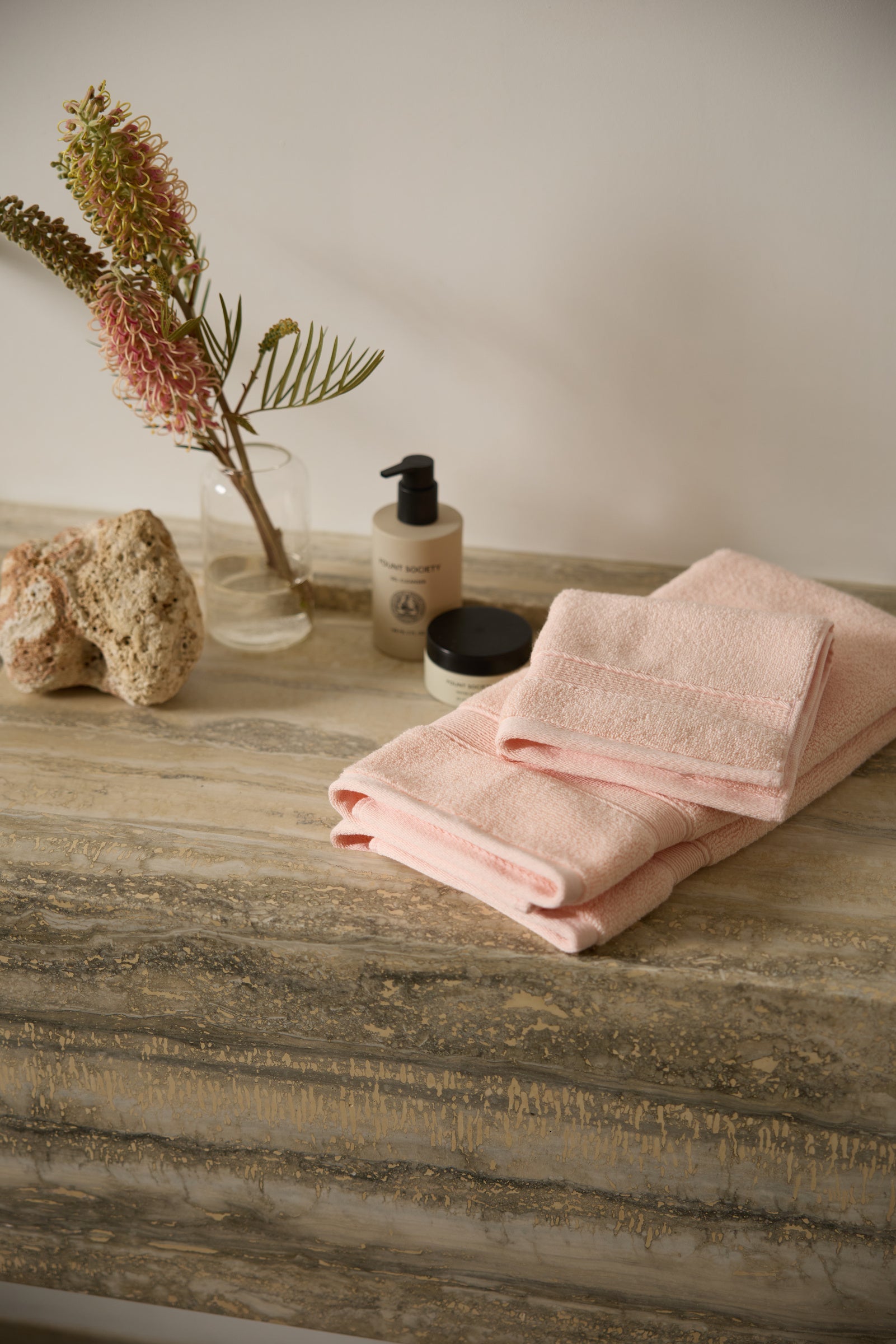 Pink towels on a textured stone counter with a bottle of lotion, a small jar, a rock, and a clear vase holding a branch with pink flowers and green foliage, set against a light wall.