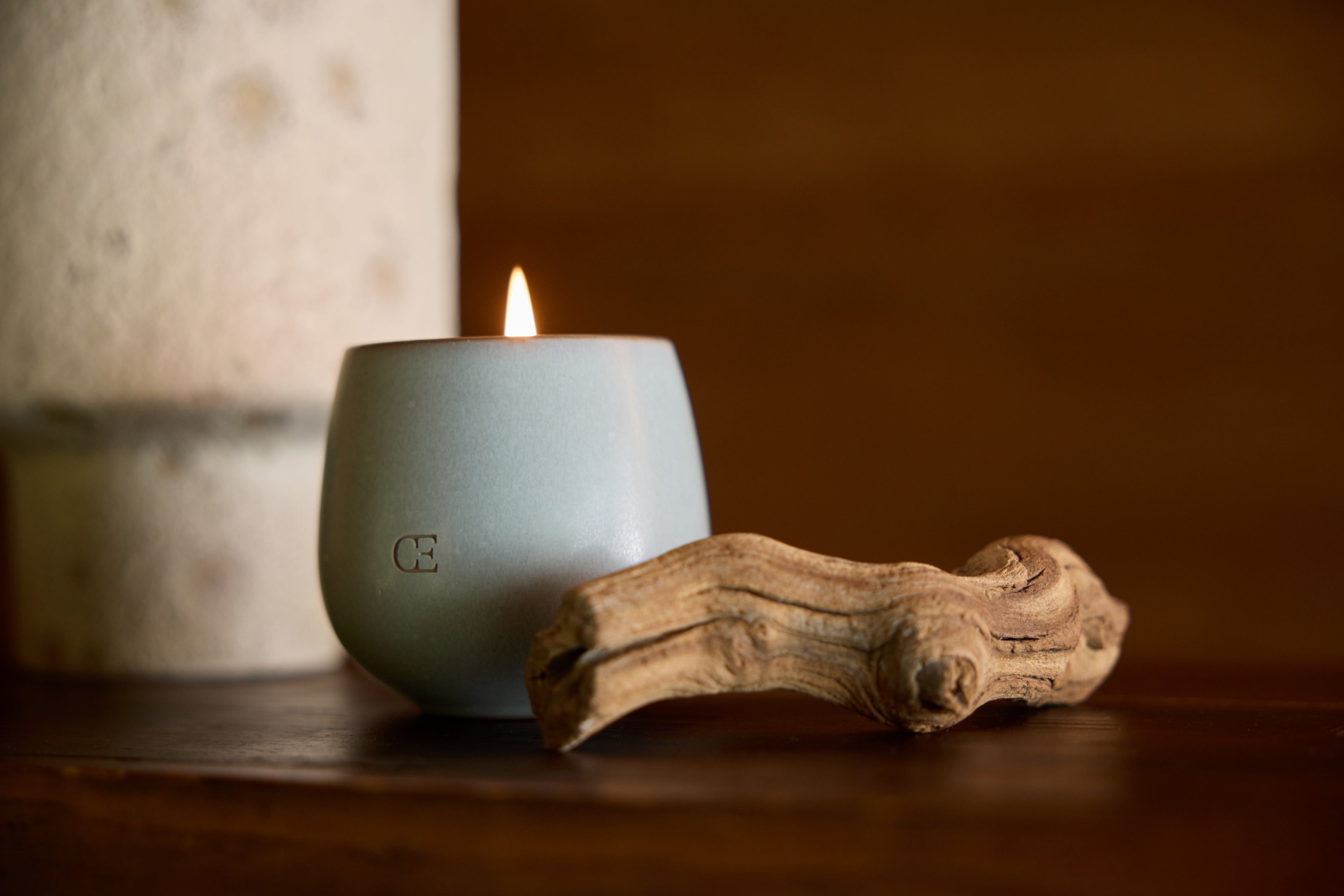 A lit candle in a smooth, blue holder sits on a wooden surface next to a piece of driftwood. A textured, cylindrical object is in the background. The setting is calm and warm.