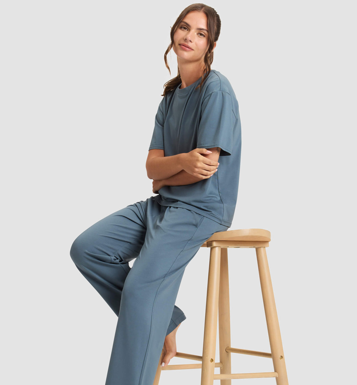 Woman with light skin and brown hair, wearing a loose blue outfit, sits barefoot on a wooden stool against a plain light background, smiling slightly with arms folded.