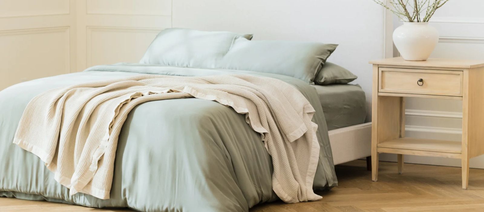 A neatly made bed with pale green bedding and a beige blanket draped over it, next to a light wooden nightstand with a white vase holding branches. The room has light walls and wooden flooring.