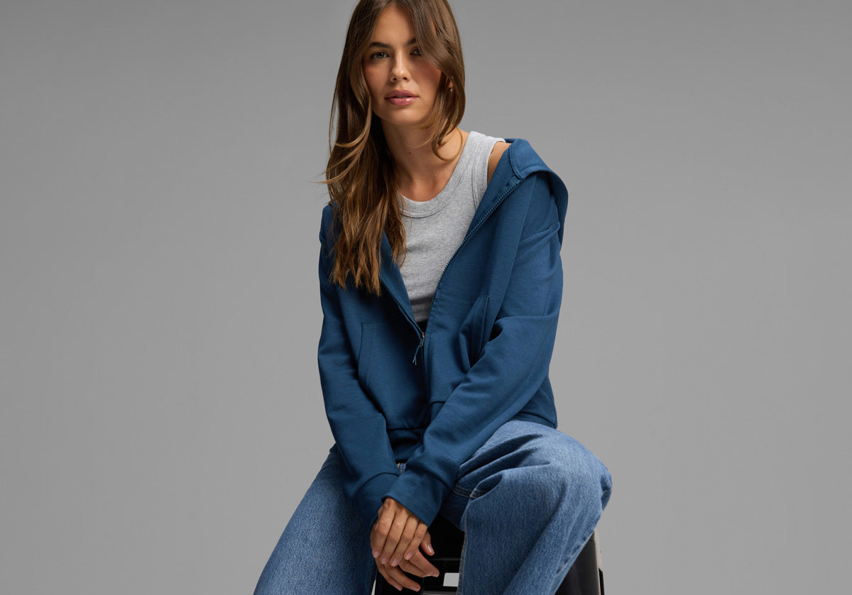 A woman with long brown hair sits on a stool, wearing a blue zip-up hoodie over a gray T-shirt and blue jeans, against a plain gray background.