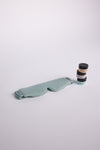 A light green sleep mask from the "Rest & Reset Bundle" by Cozy Earth is on a white surface, next to a pyramid stack of three "Plant Remedy" jars, each featuring a unique colored lid.