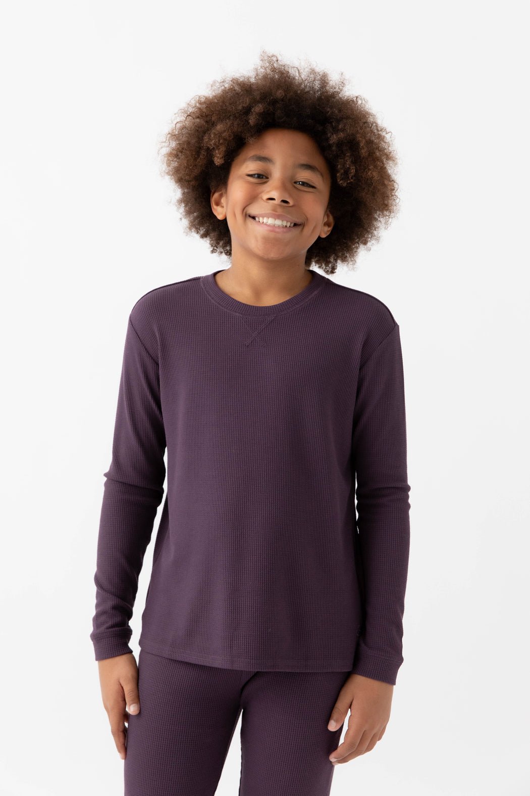 A smiling child with curly hair stands against a white background, wearing the Cozy Earth Children's Bamboo Waffle Knit Base Layer Long Sleeve in dark purple.|Color:Plum