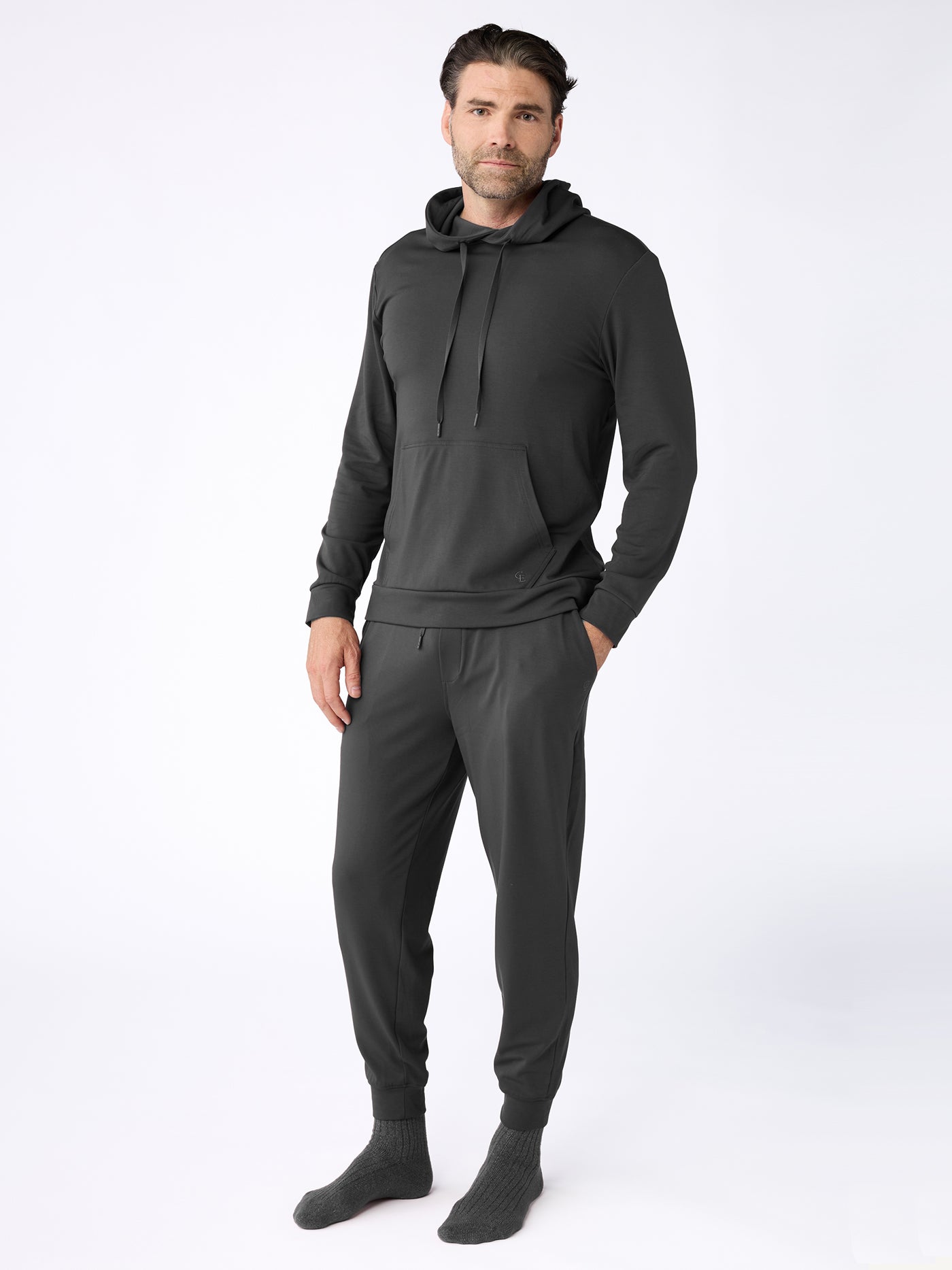 A man wears the Cozy Earth Men's Brushed Bamboo Hoodie & Jogger Set in dark gray, with his hands in his pockets and gray socks, posing against a plain white background. |Color:Charcoal