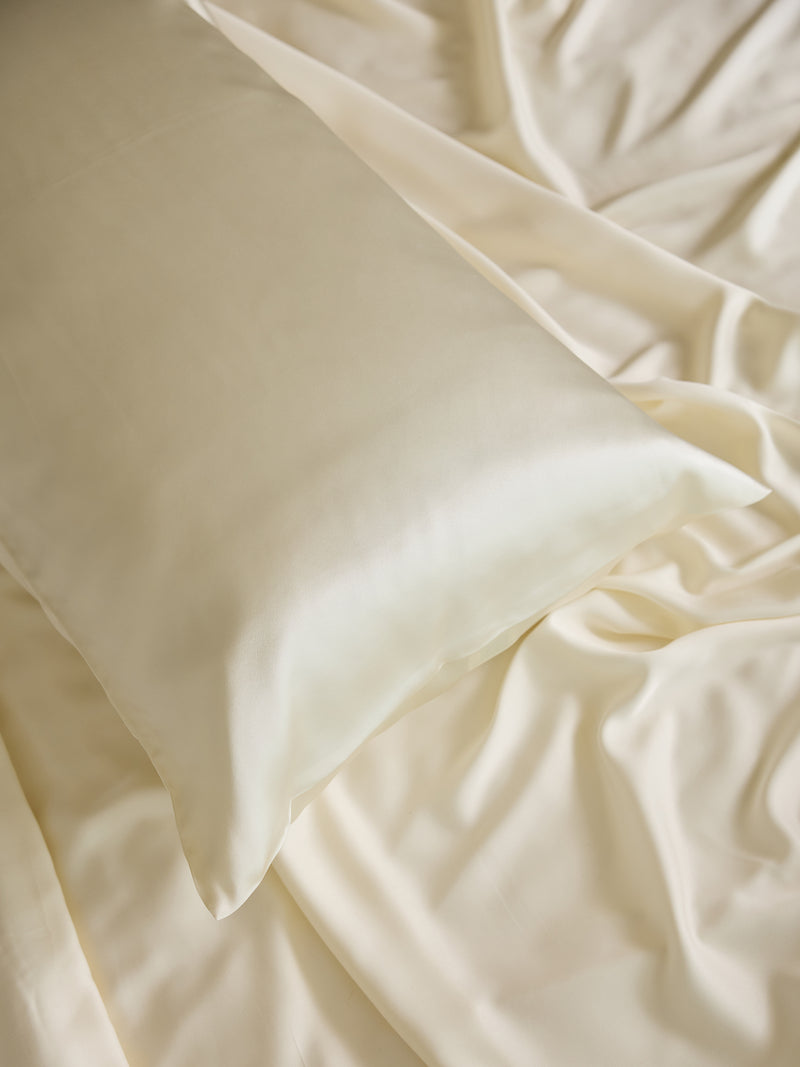 A close-up of a silky, cream-colored Silk Pillowcase by Cozy Earth on matching satin sheets. The fabric has a smooth and shiny texture, creating a luxurious and elegant appearance.