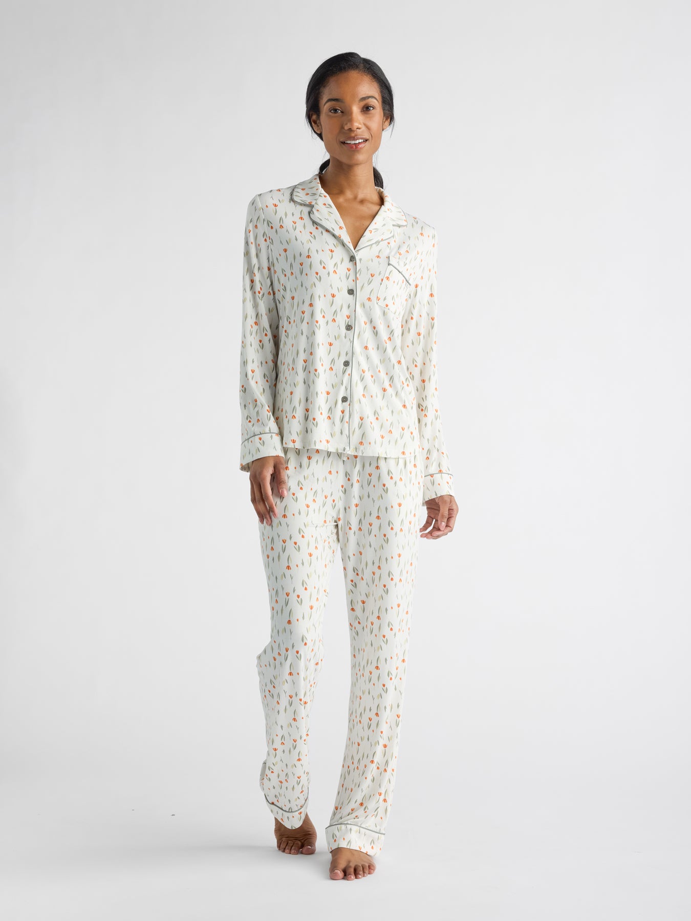 Wearing HIDE's Women's Bamboo Stretch-Knit Long Sleeve Pajama Top, a person stands barefoot on a white surface, clad in matching pajamas with a light background and small colorful patterns, smiling as they look forward.