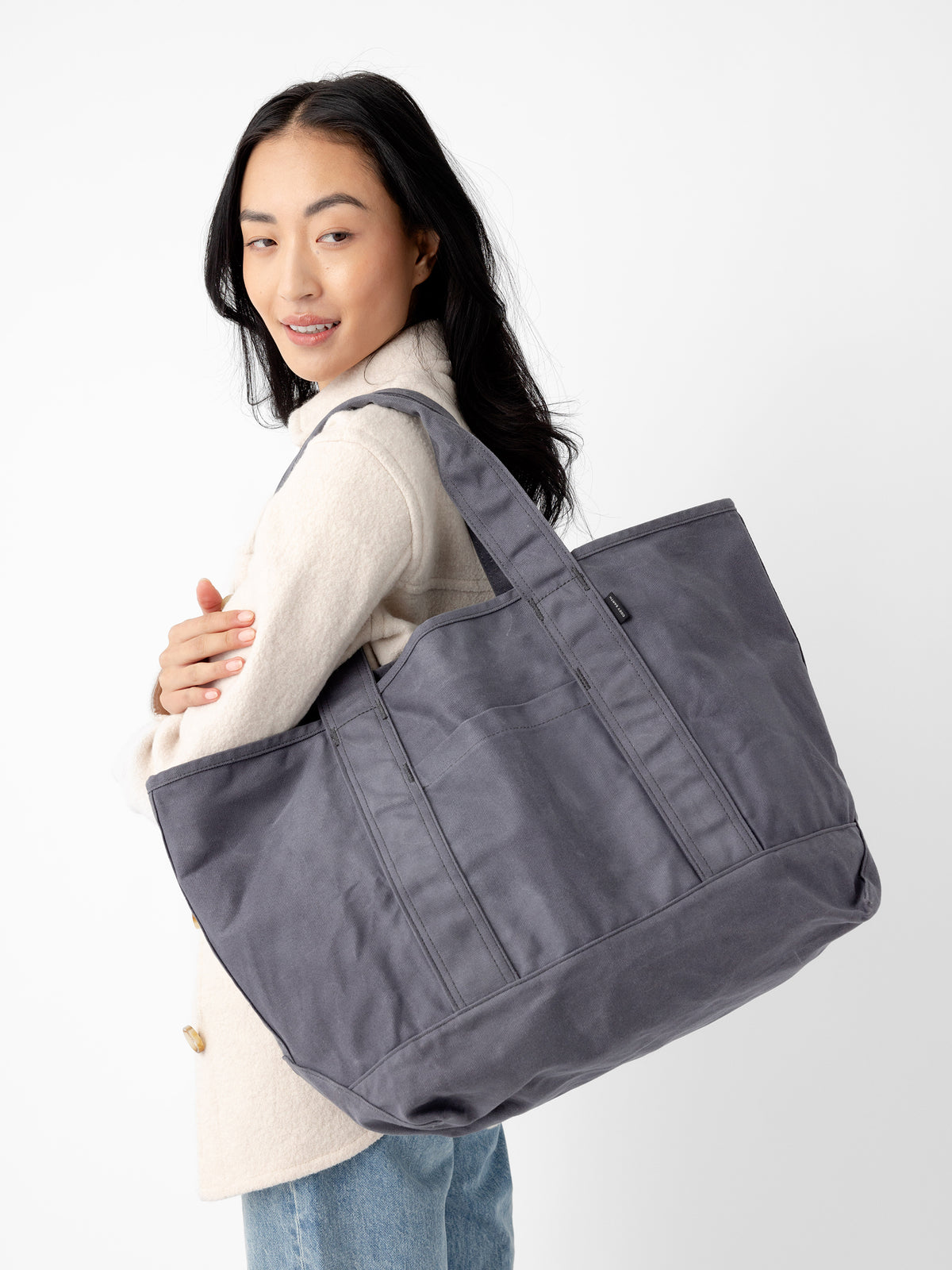A person with long dark hair is wearing a light-colored coat and blue jeans. They have a large Cozy Earth Waxed Canvas Tote over their shoulder. The background is plain white. |Color:Washed Navy