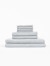 The Cozy Earth Nantucket Bath Towel Set features light grey textured towels in multiple sizes, neatly stacked from largest to smallest against a plain white background.|Color:Heathered Harbor Mist