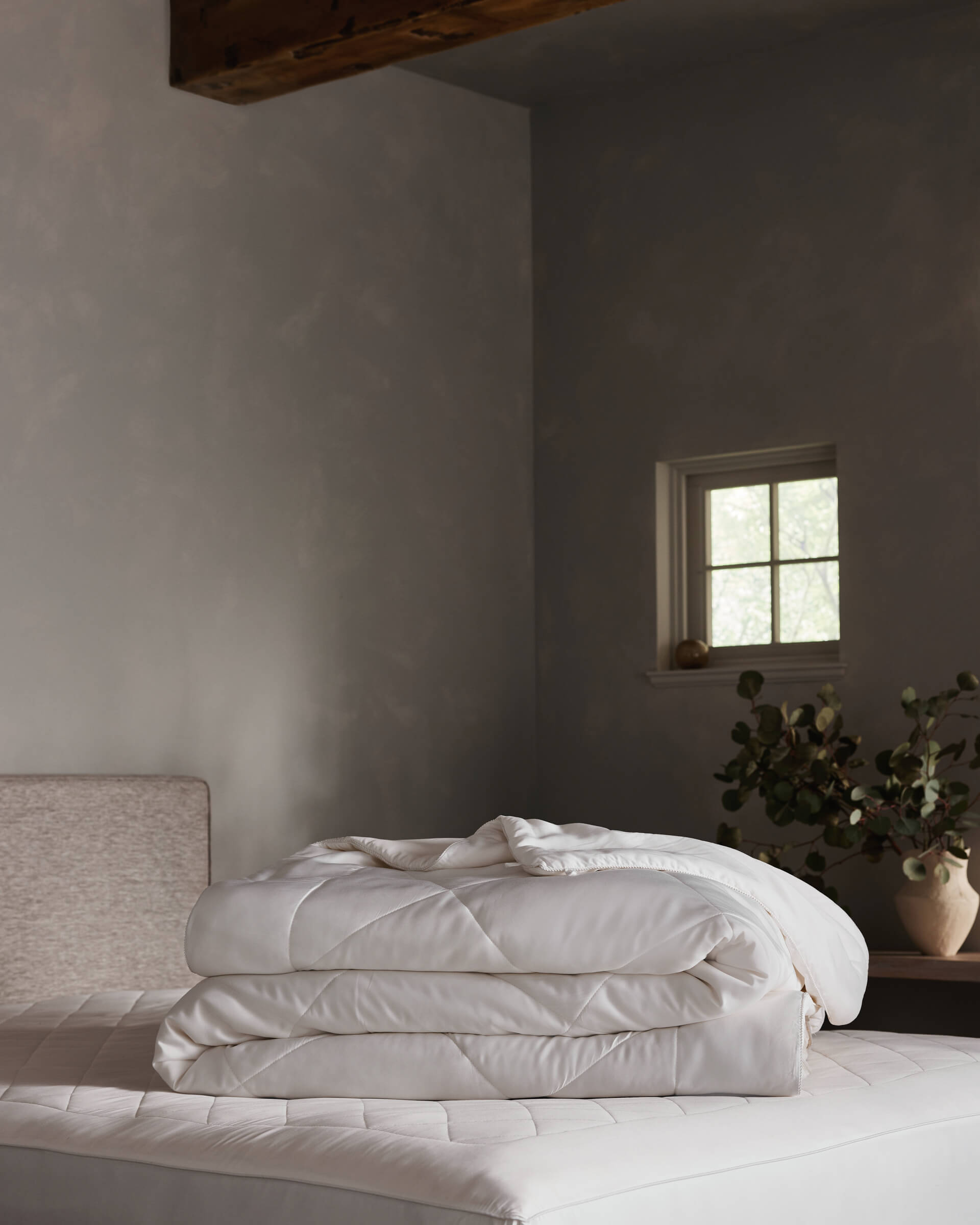 The Soft-Wash Cotton Bedding Deluxe Bundle by Cozy Earth, neatly folded, rests on a bed in a minimalist bedroom featuring light gray walls, a small window, and a wooden nightstand with a vase of green branches.