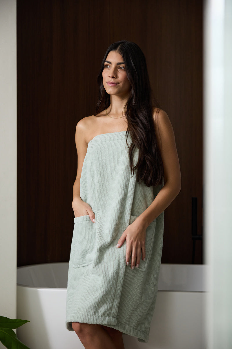 Someone wears Cozy Earth's Ribbed Terry Bath Wrap in beige, featuring ribbed fabric and large front pockets—one hand rests inside a pocket with the Cozy Earth label showing.|Color:Petal