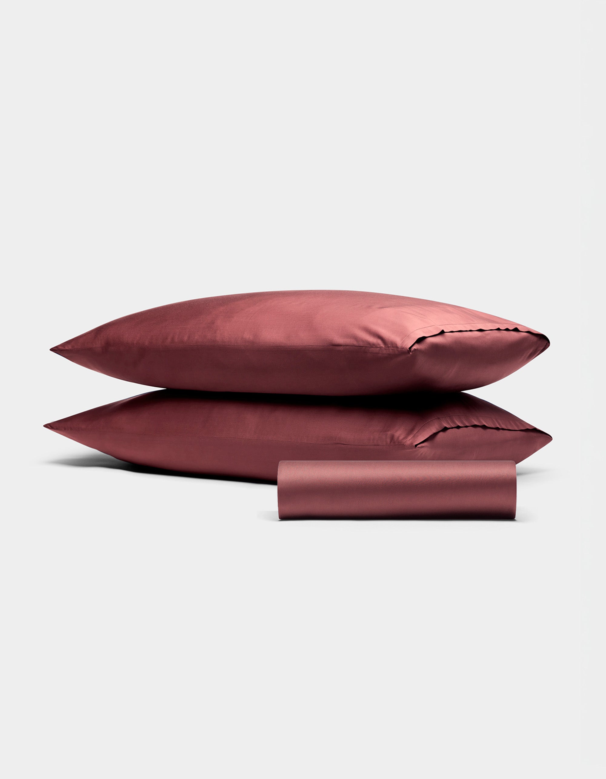 Two stacked, burgundy Bamboo Bedding Basic Bundle pillowcases by Cozy Earth with a matching rolled-up sheet, all set against a plain light gray background.