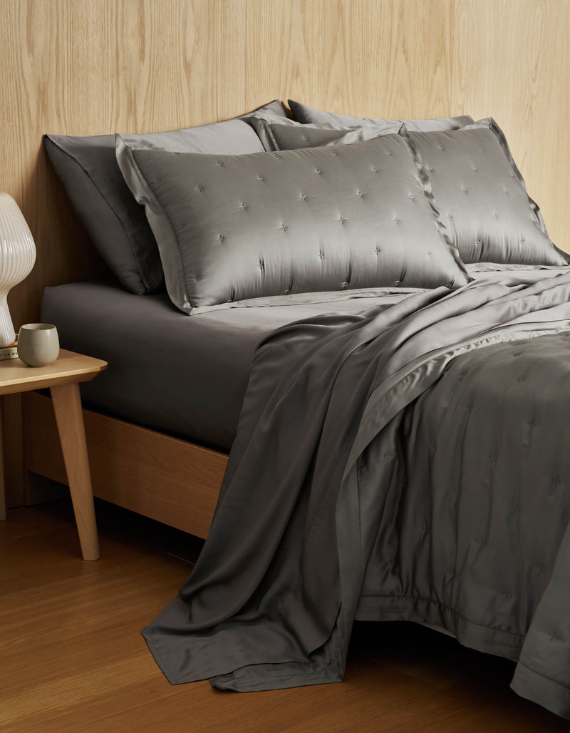 A neatly made bed features the Cozy Earth Bamboo Quilt Bundle—gray, silky bedding with embroidered pillows—beside a wooden nightstand holding a white lamp and mug, set against a light wood-paneled wall.