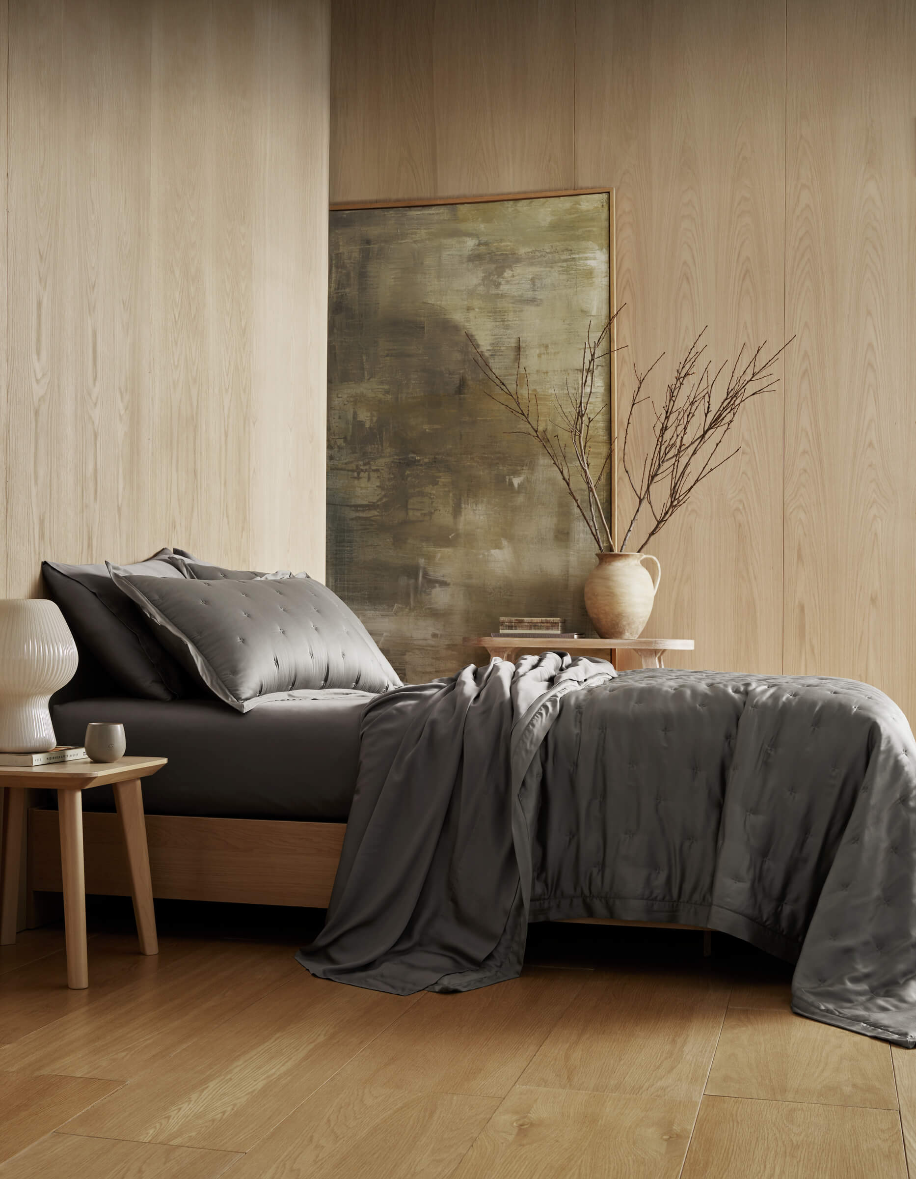 A modern bedroom with light wood walls and floors showcases a neatly made bed featuring the Cozy Earth Bamboo Quilt Bundle, complemented by a bedside table with a white lamp, a ceramic vase with bare branches, and a large abstract painting.|Color:Charcoal