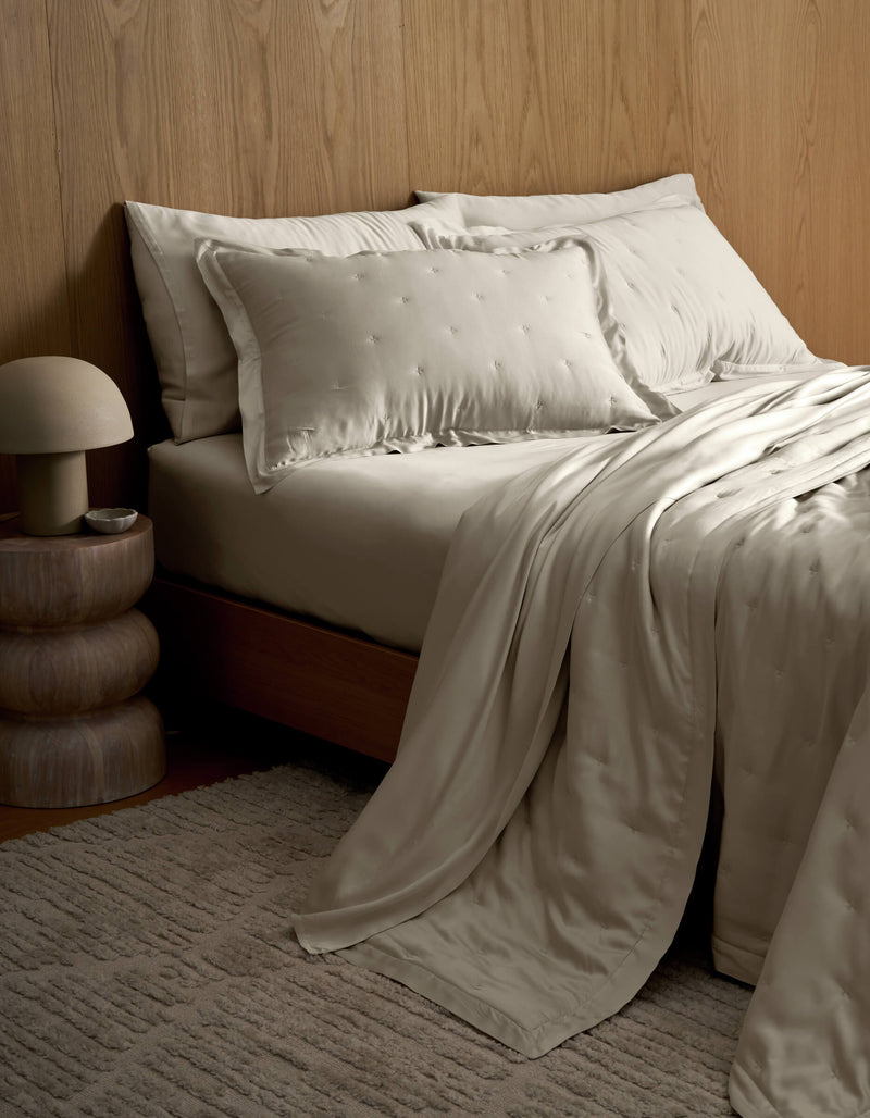 A neatly made bed featuring Cozy Earth's Bamboo Quilt with satin pillows sits beside a wooden nightstand with a round lamp, all set against a wood-paneled wall and a textured beige rug.