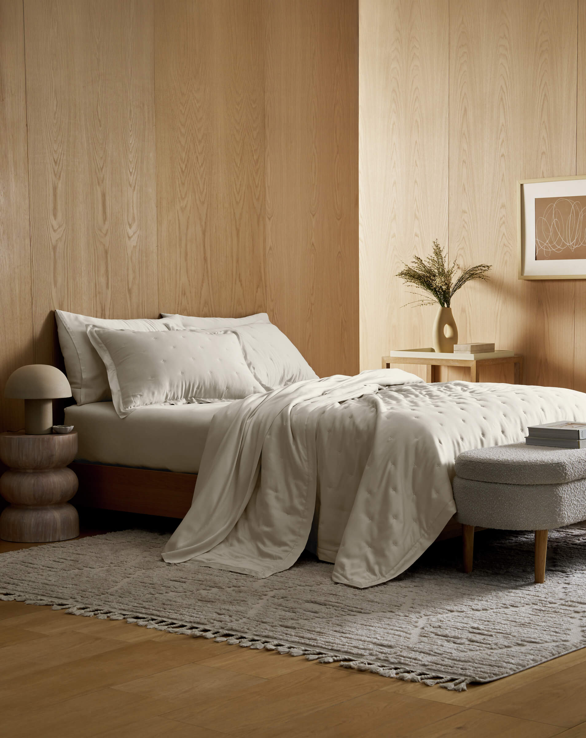 A minimalist bedroom with light wood walls, a neatly made bed featuring the Cozy Earth Bamboo Quilt Bundle, a small nightstand with a vase and dried flowers, a textured rug, round stool, and framed art on the wall.