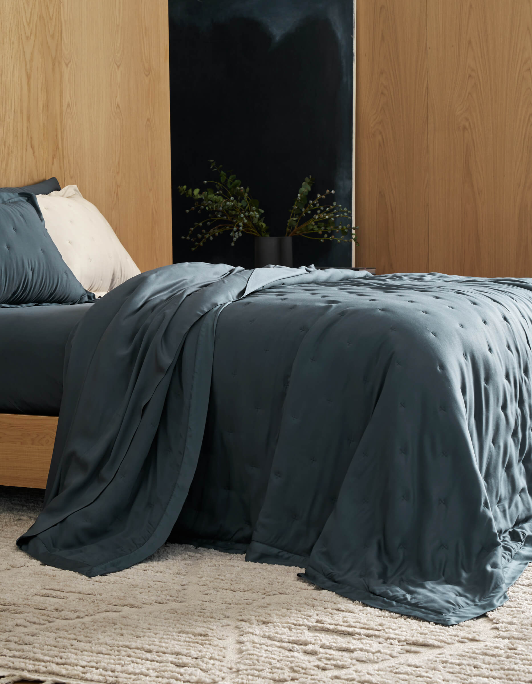 A modern bedroom features the Cozy Earth Bamboo Quilt Bundle on the bed, its dark blue texture matching the pillow. Light wood walls, a beige rug, and a black vase with green foliage contrast with the room’s neutral decor.|Color:Pacific Blue